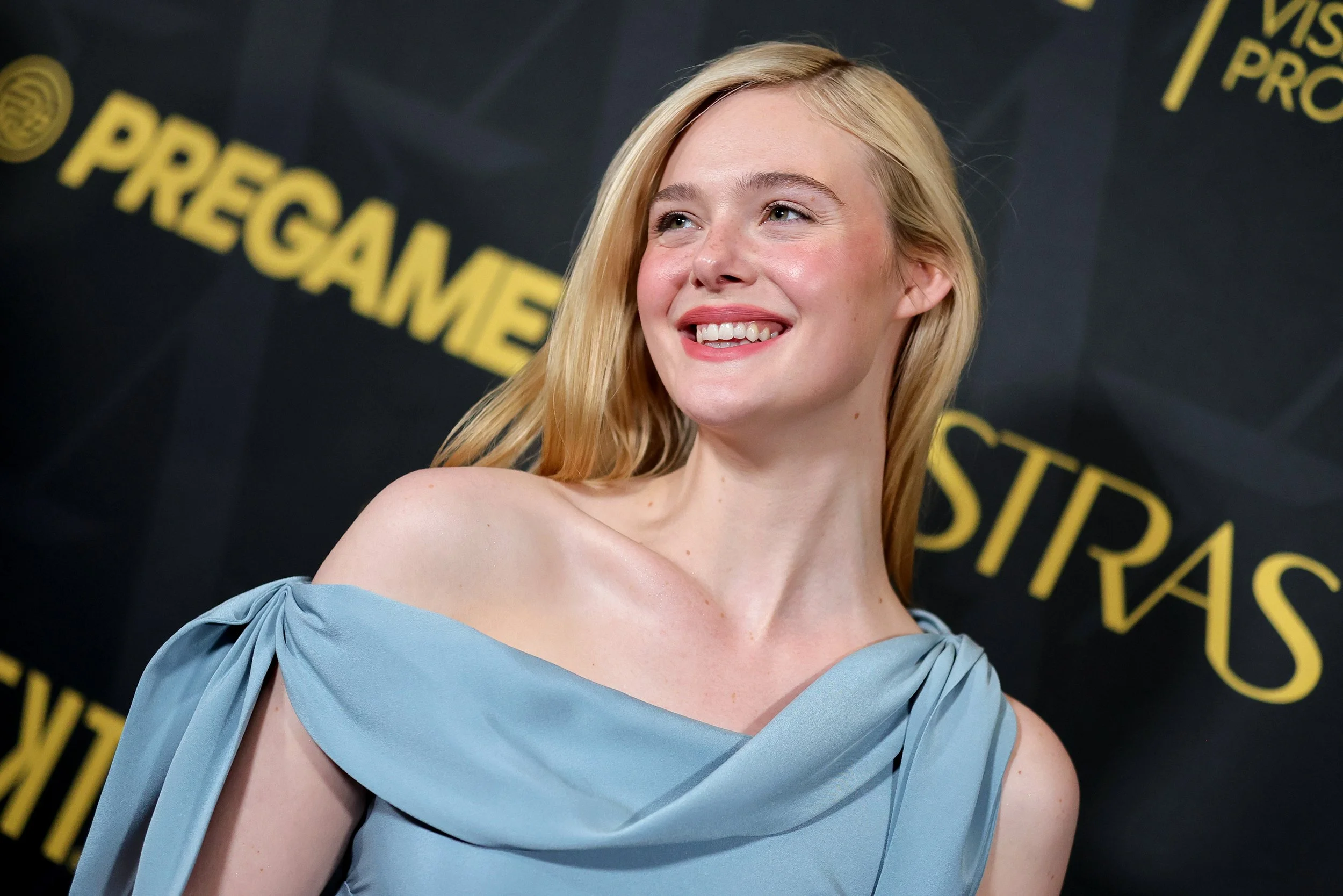 A blonde woman smiling, wearing a light blue off-shoulder dress, standing in front of a black backdrop with yellow text that reads 'PREGAMES' and 'STRAS'.