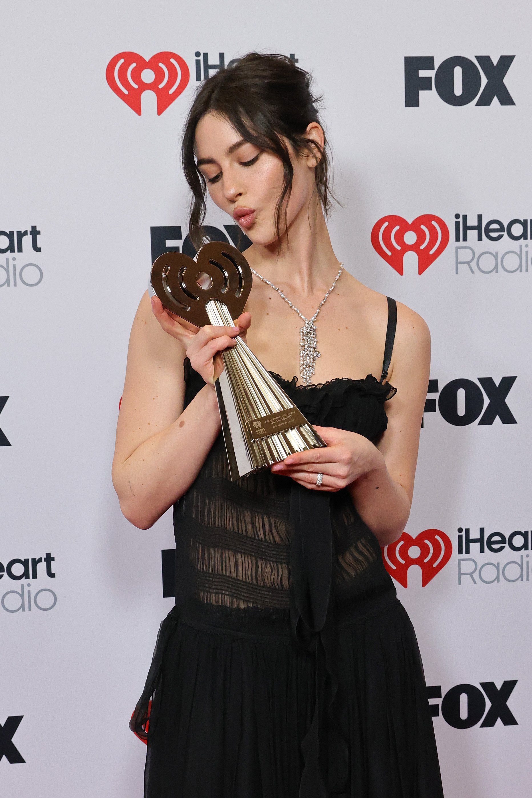 A woman in a black dress holding an iHeartRadio award at an event with a backdrop featuring the iHeartRadio and FOX logos.