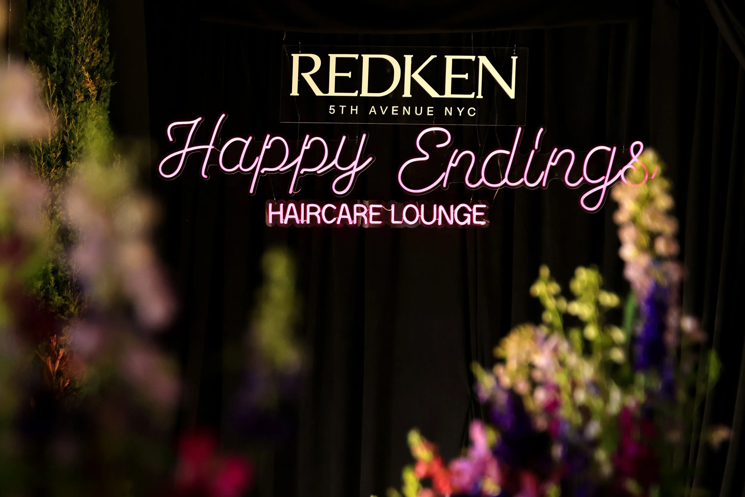 Neon sign with 'Happy Endings Haircare Lounge' and 'Redken 5th Avenue NYC' against a black background, surrounded by blurred colorful flowers.
