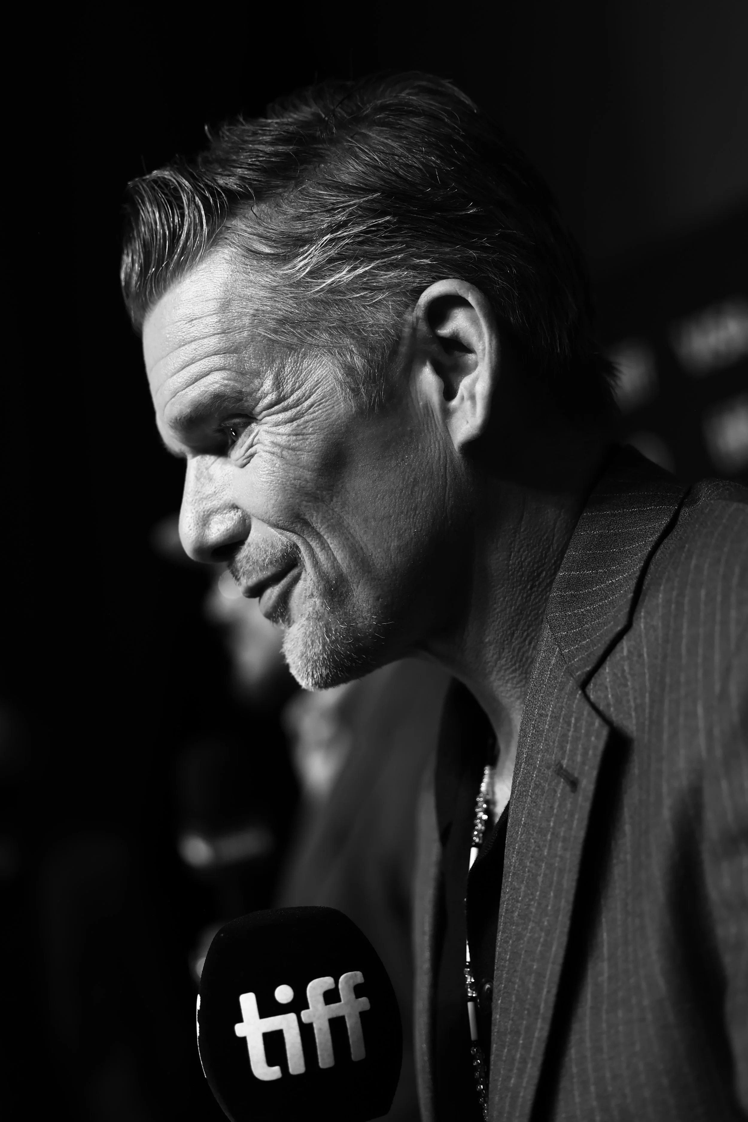 Black and white photo of a man in a suit speaking into a microphone with 'tiff' logo, with a side profile, hair slicked back, in a dimly lit environment.