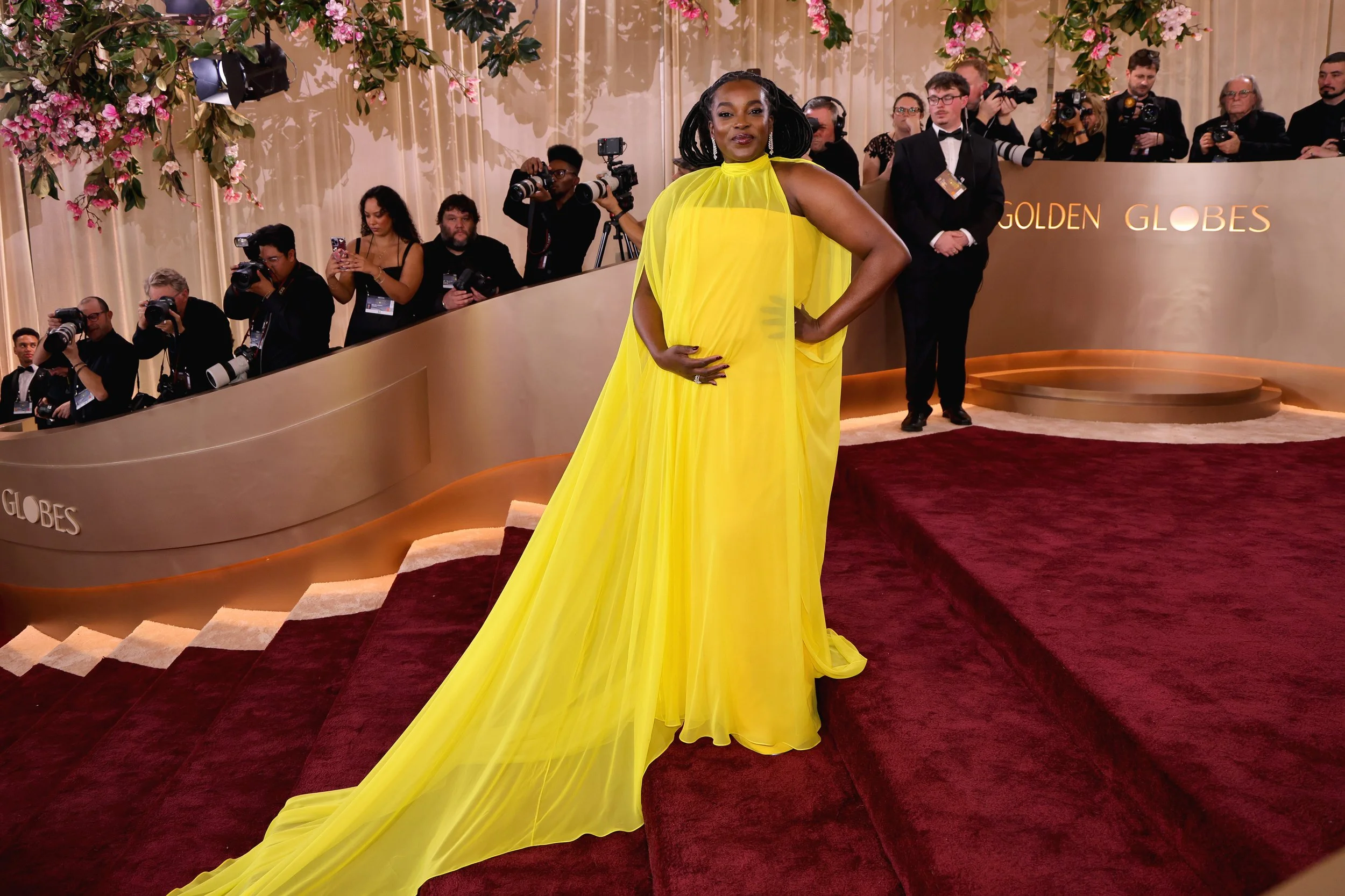 A woman in a bright yellow gown standing on a red carpet at an event with photographers and attendees in the background.