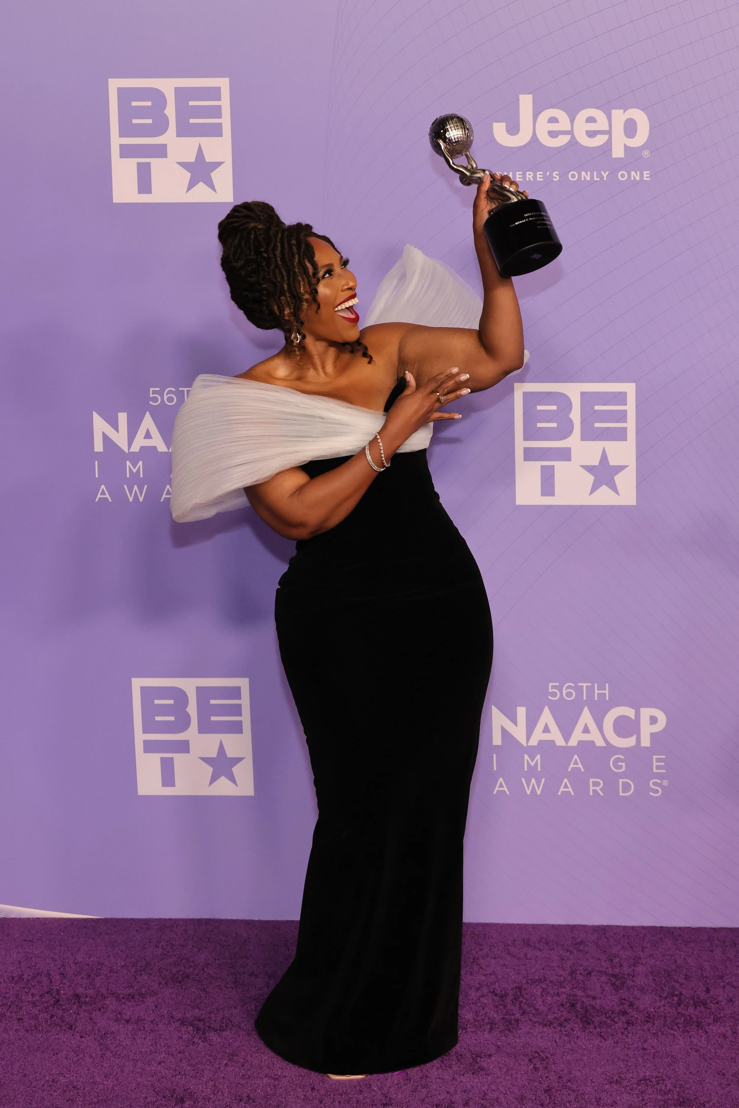 A woman in an elegant black dress with a white pleated shoulder detail, celebrating with a trophy at the NAACP Image Awards, standing on purple carpet in front of a purple backdrop with event logos.