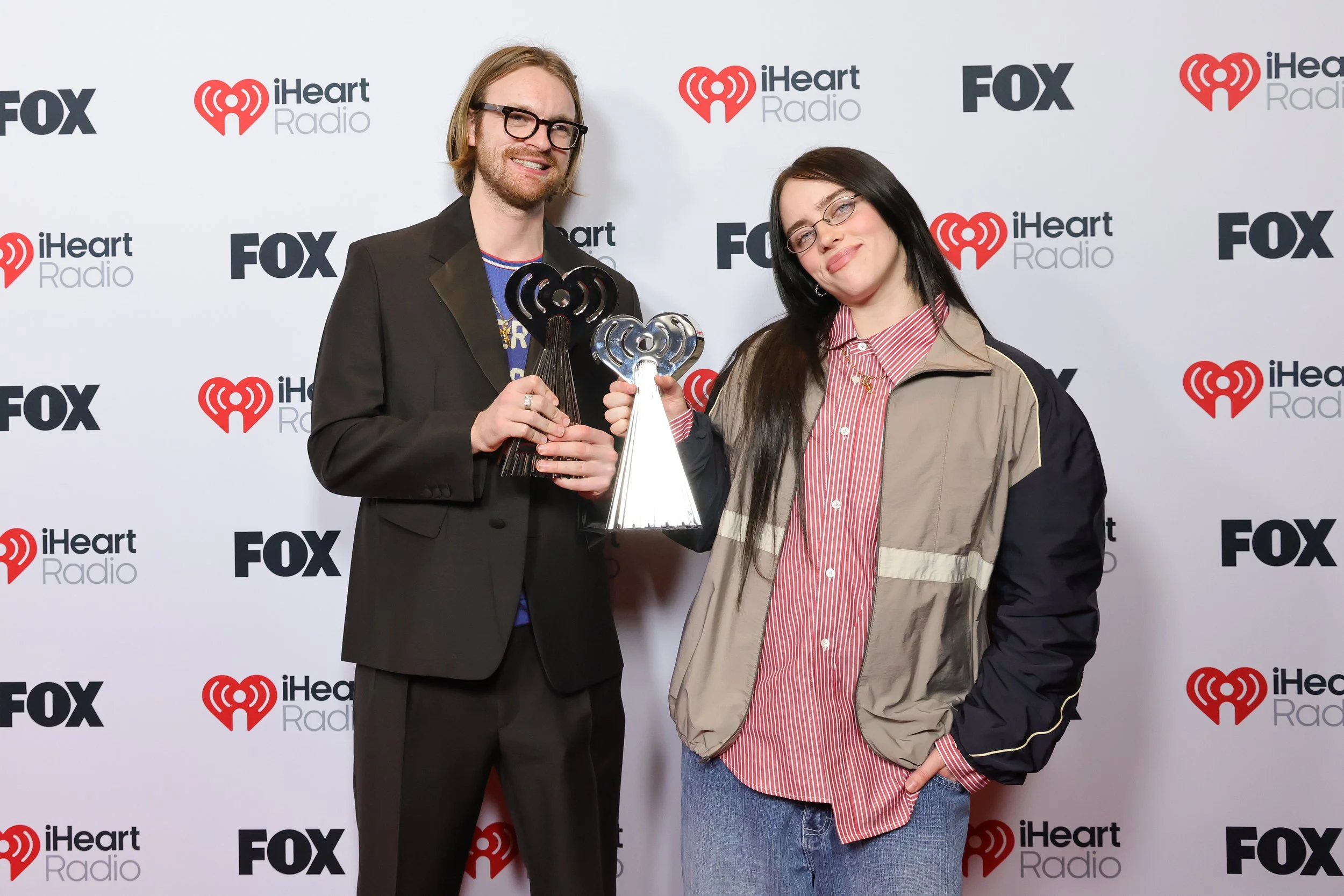 Two people, a man and a woman, standing in front of a backdrop with iHeartRadio and FOX logos, holding award trophies and smiling.