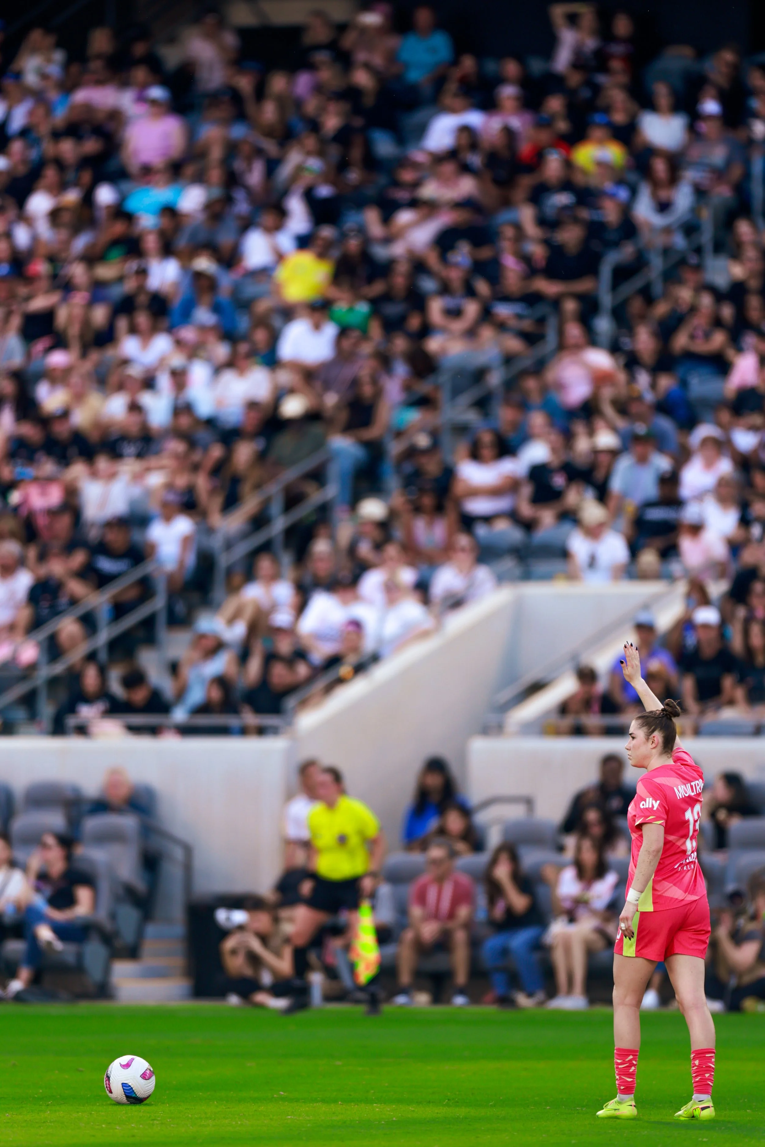A woman in a pink soccer uniform preparing to take a penalty kick on a green field, with a large crowd of spectators in the stands behind her.