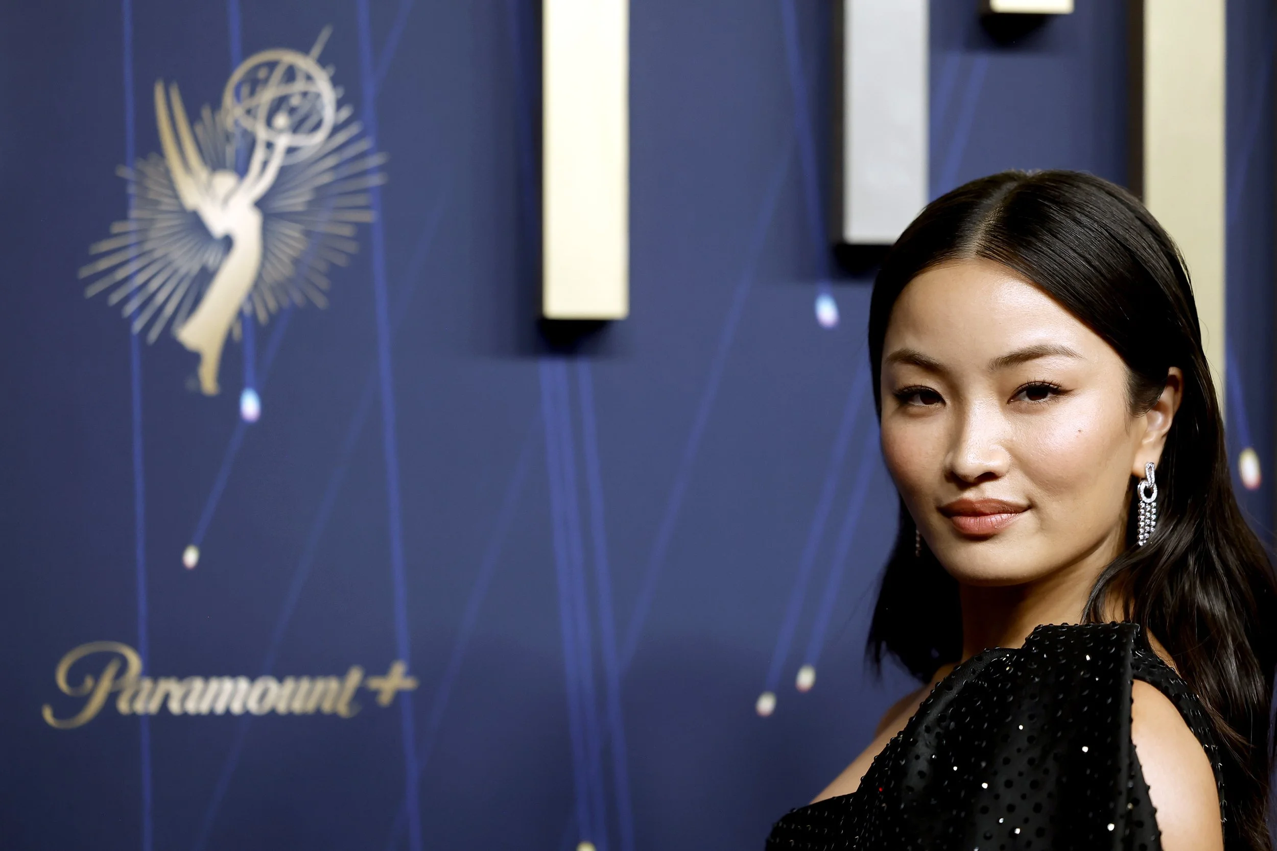 A woman with dark hair in an elegant black dress with sparkles, wearing earrings, standing in front of a blue backdrop with a gold logo of a winged figure and the text 'Paramount+'.