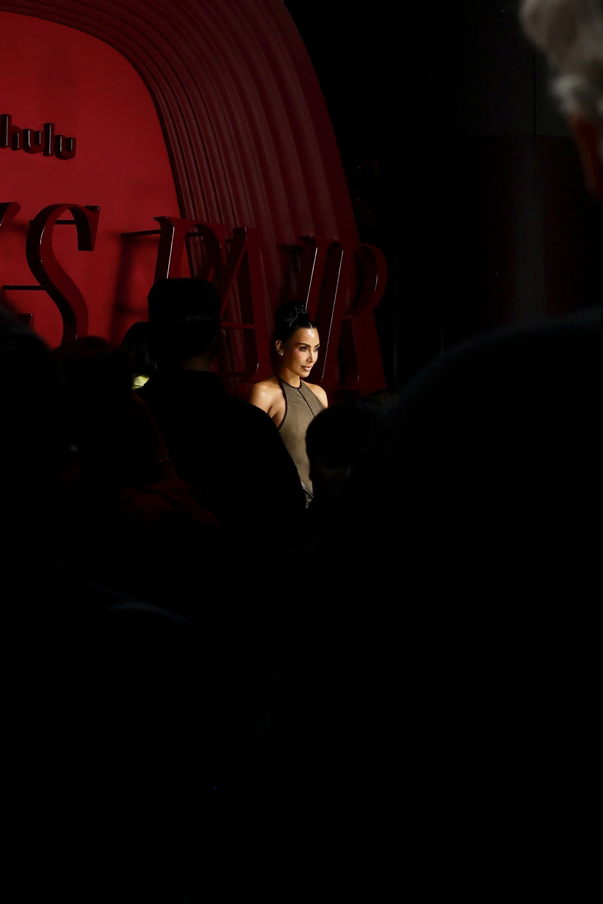 A woman on stage at a Hulu event, with an audience in front of her, red backdrop with Hulu logo, stage lighting highlighting her.