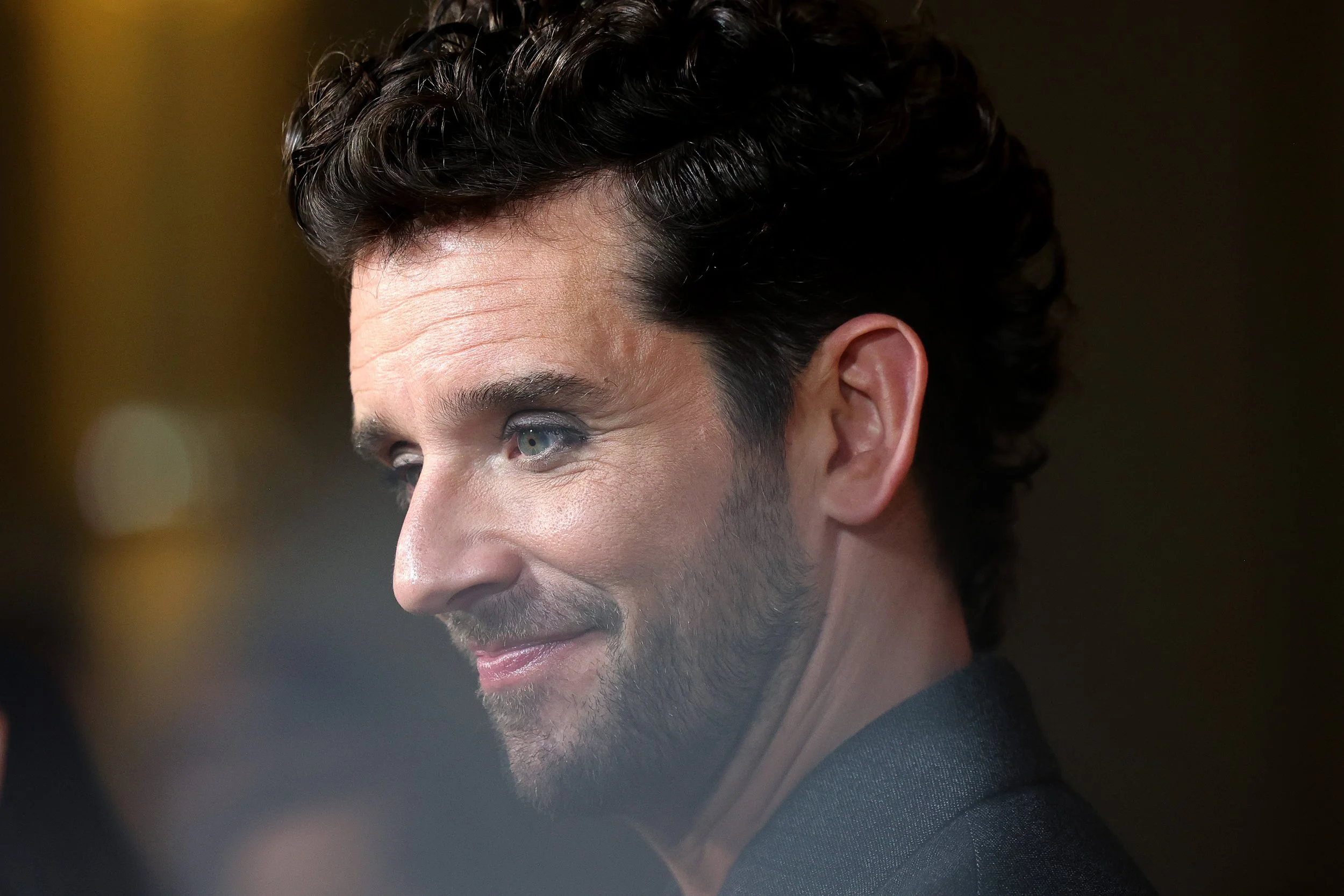 Close-up of a man with dark, curly hair and a beard smiling, wearing a dark blazer, with a blurred background.
