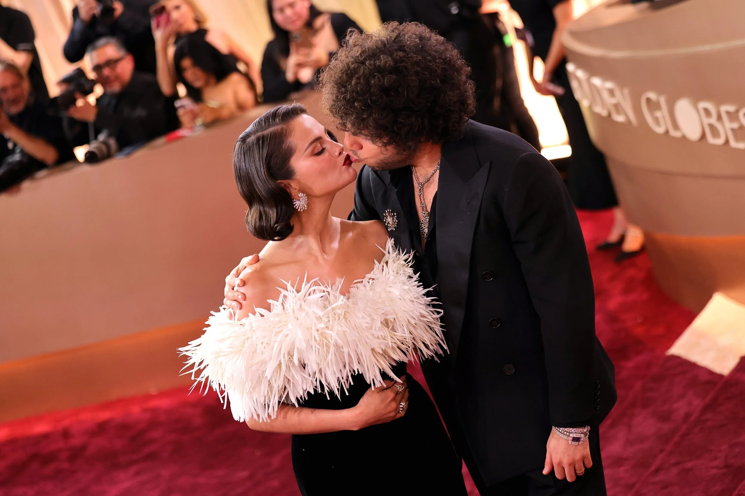 A man and woman kissing at an awards ceremony, with a crowd of onlookers in the background.