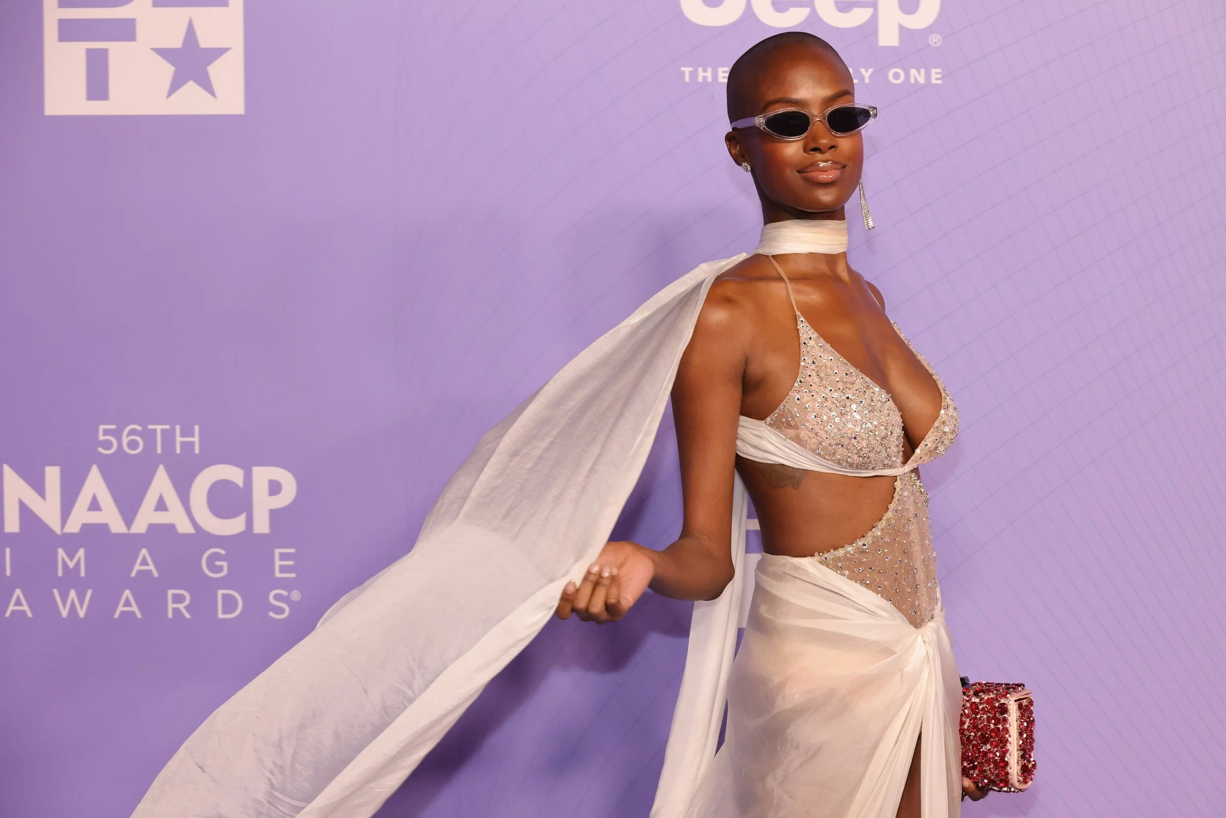Woman in a sparkly beige dress with a cutout, wearing sunglasses, holding a red clutch, and posing at the NAACP Image Awards.