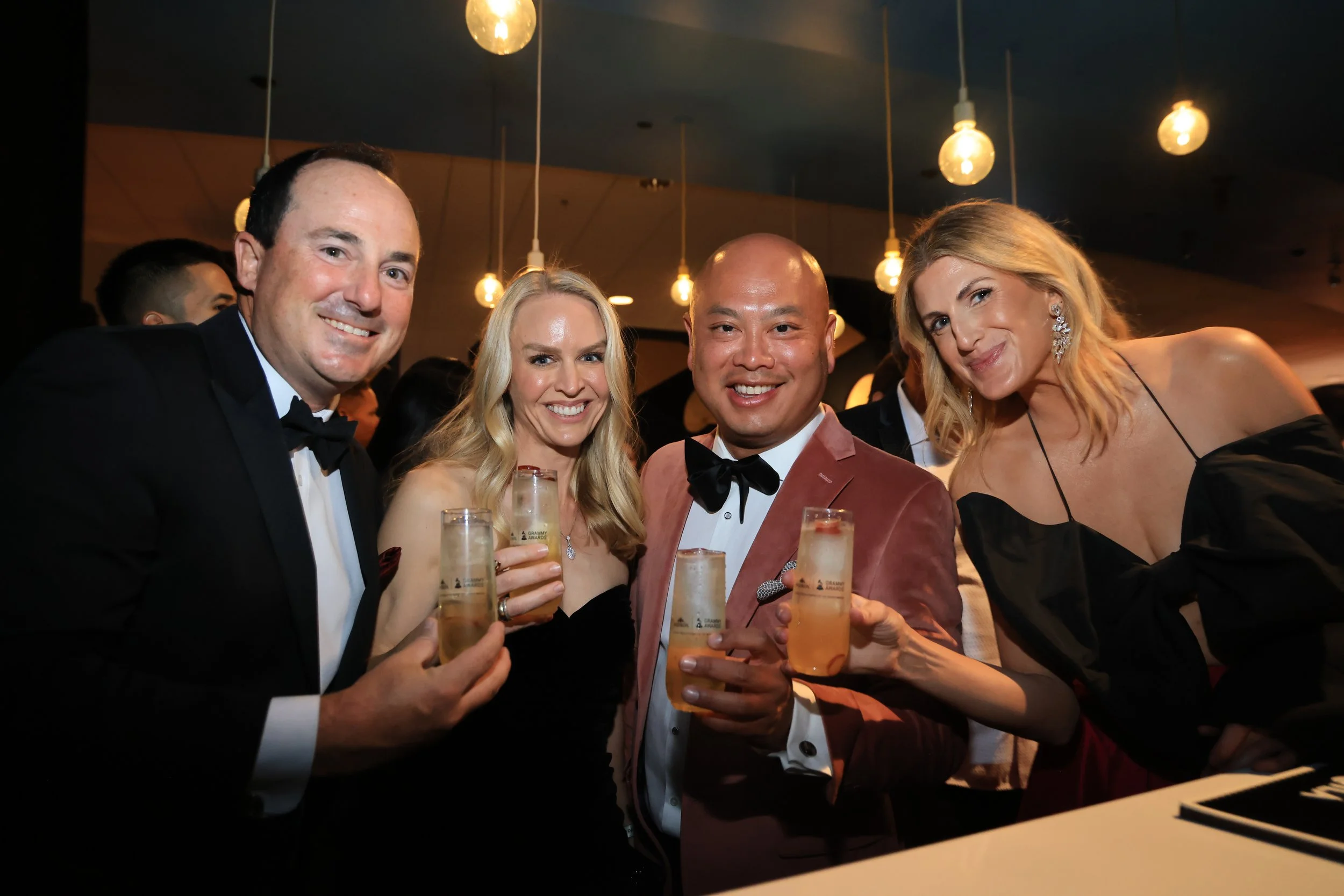 Four people in formal attire holding glasses of champagne at a party or celebration, smiling at the camera.