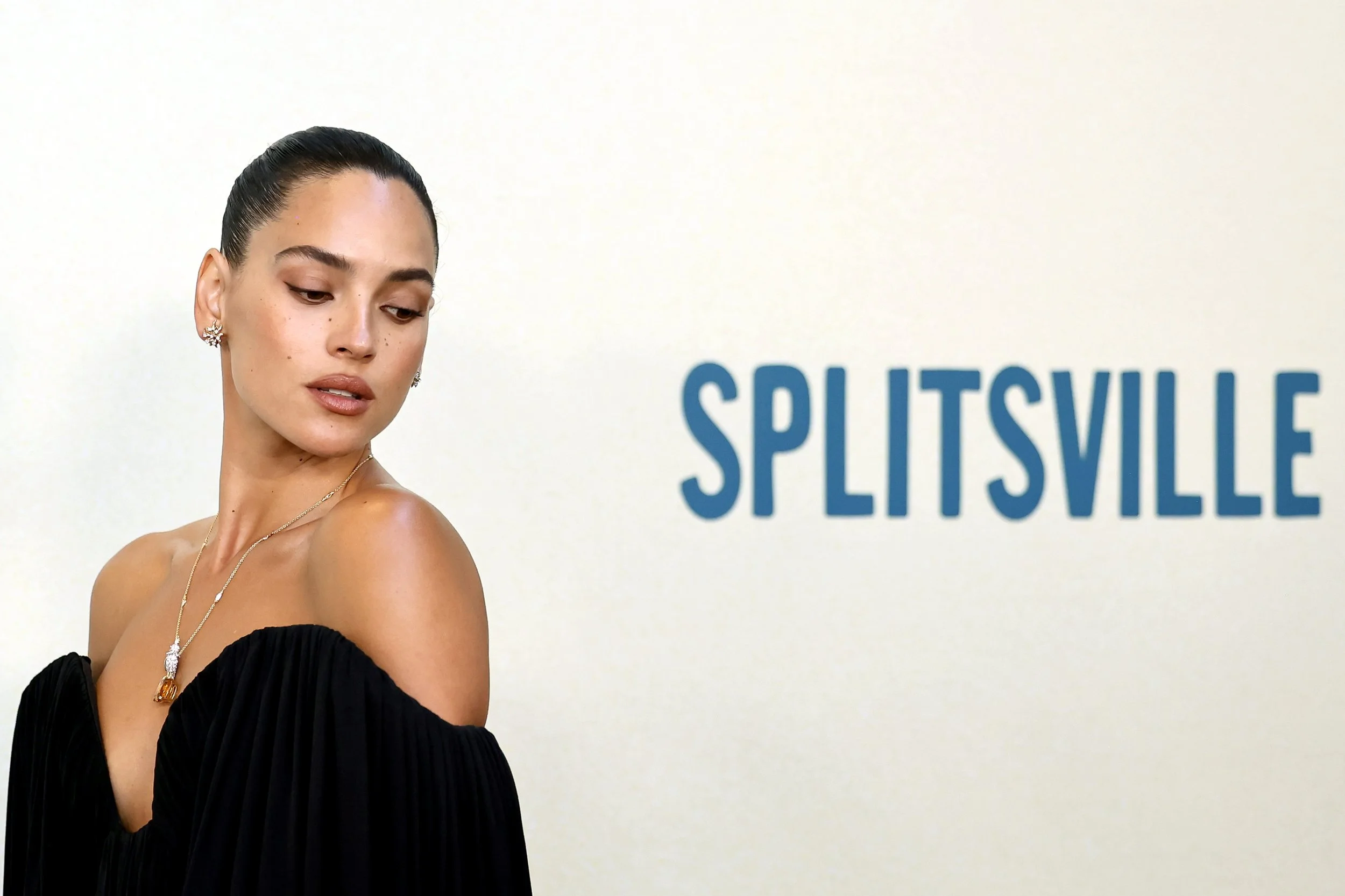A woman with dark hair in a bun wearing an off-the-shoulder black dress and jewelry, standing next to a wall with the blue text 'SPLITSVILLE'.