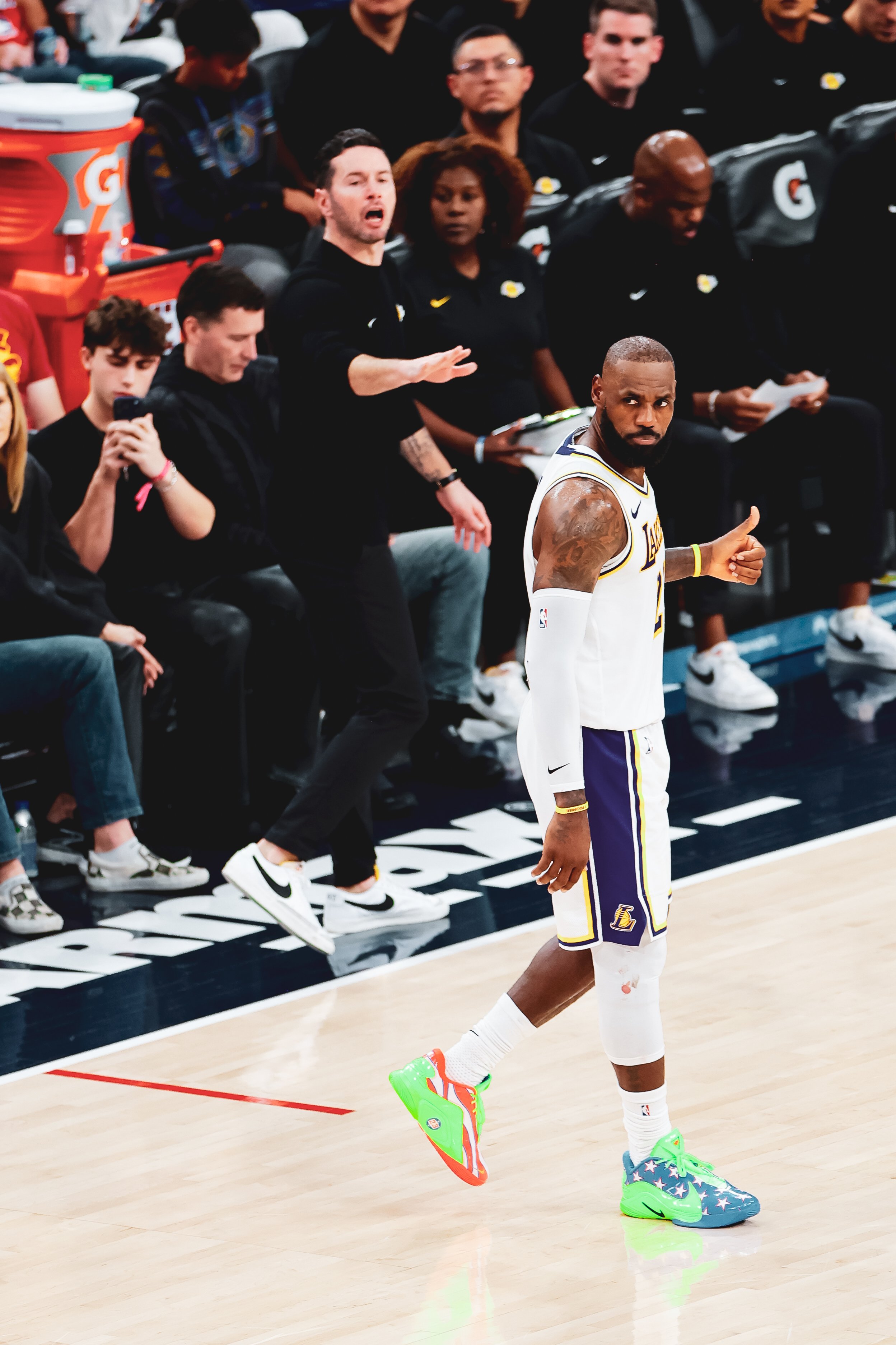 Basketball player in a Lakers uniform giving a thumbs up on the court during a game, with fans and staff sitting courtside in the background.