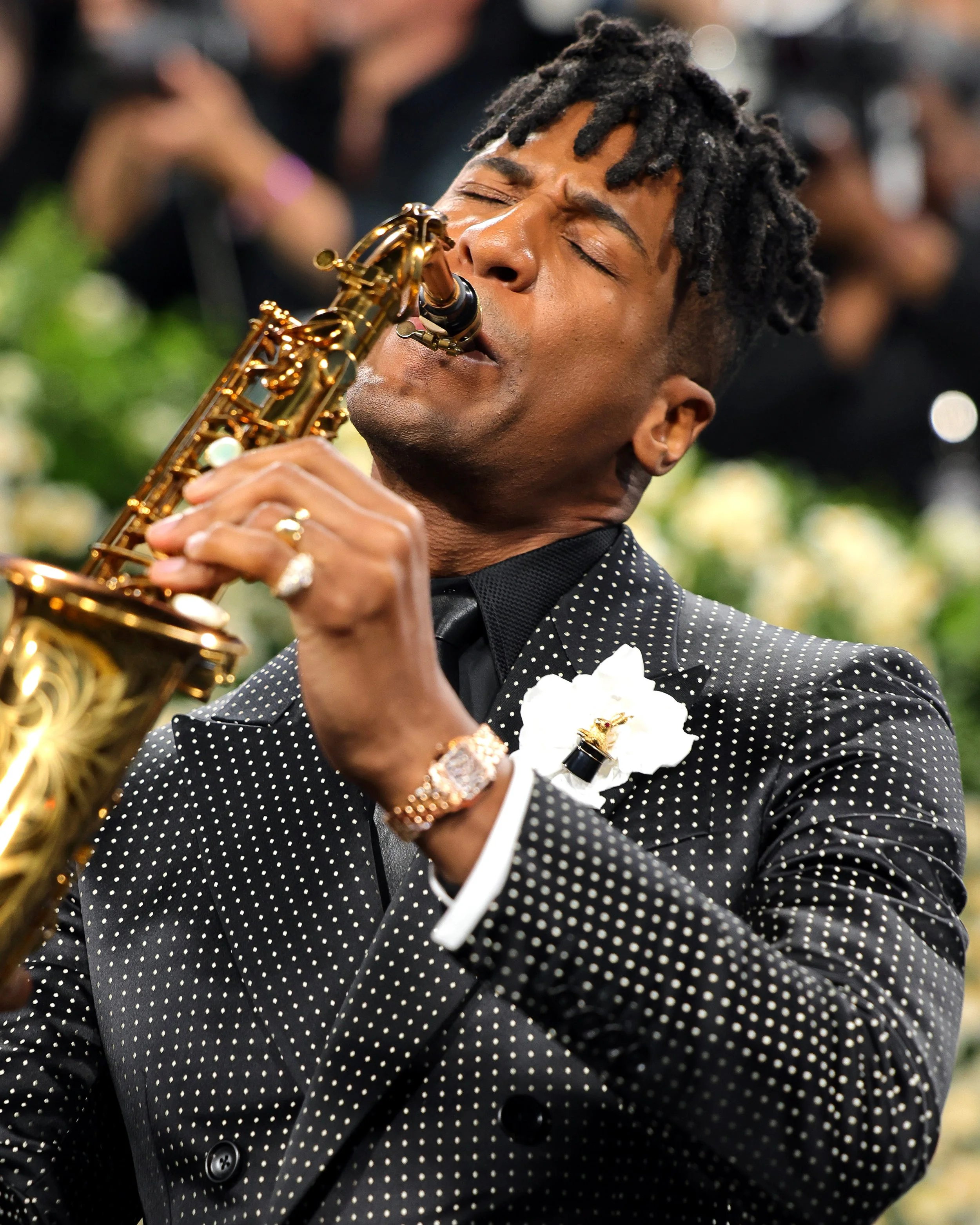Man in a dark polka dot suit playing a gold saxophone with closed eyes at an event, with a white flower pin on his lapel and a background of green and white flowers.