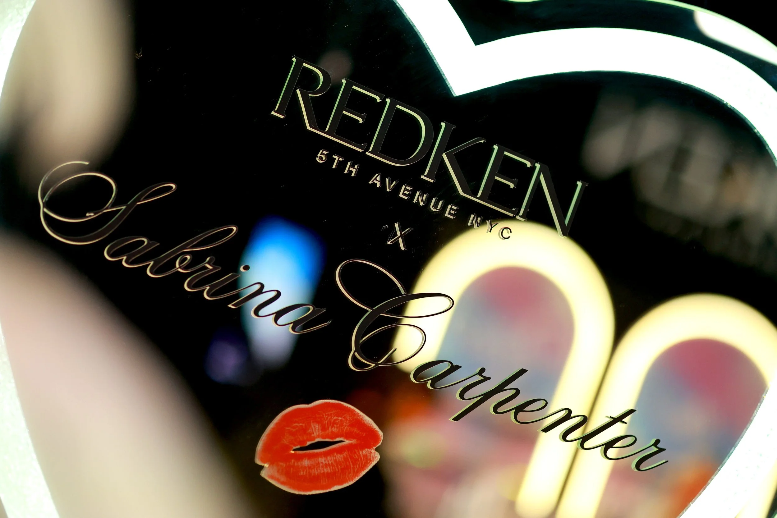 A neon sign reads 'Redken 5th Avenue NYC' with the words 'Celebrating Mother' in cursive, accompanied by a red lipstick kiss mark.
