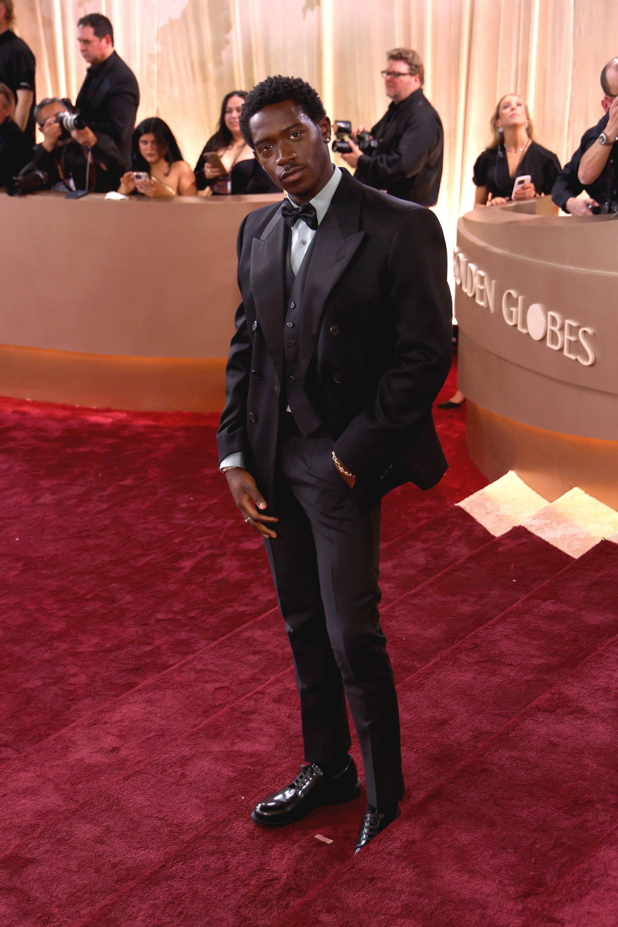 A young man in a black tuxedo with a bow tie posing on a red carpet at the Golden Globes awards ceremony.