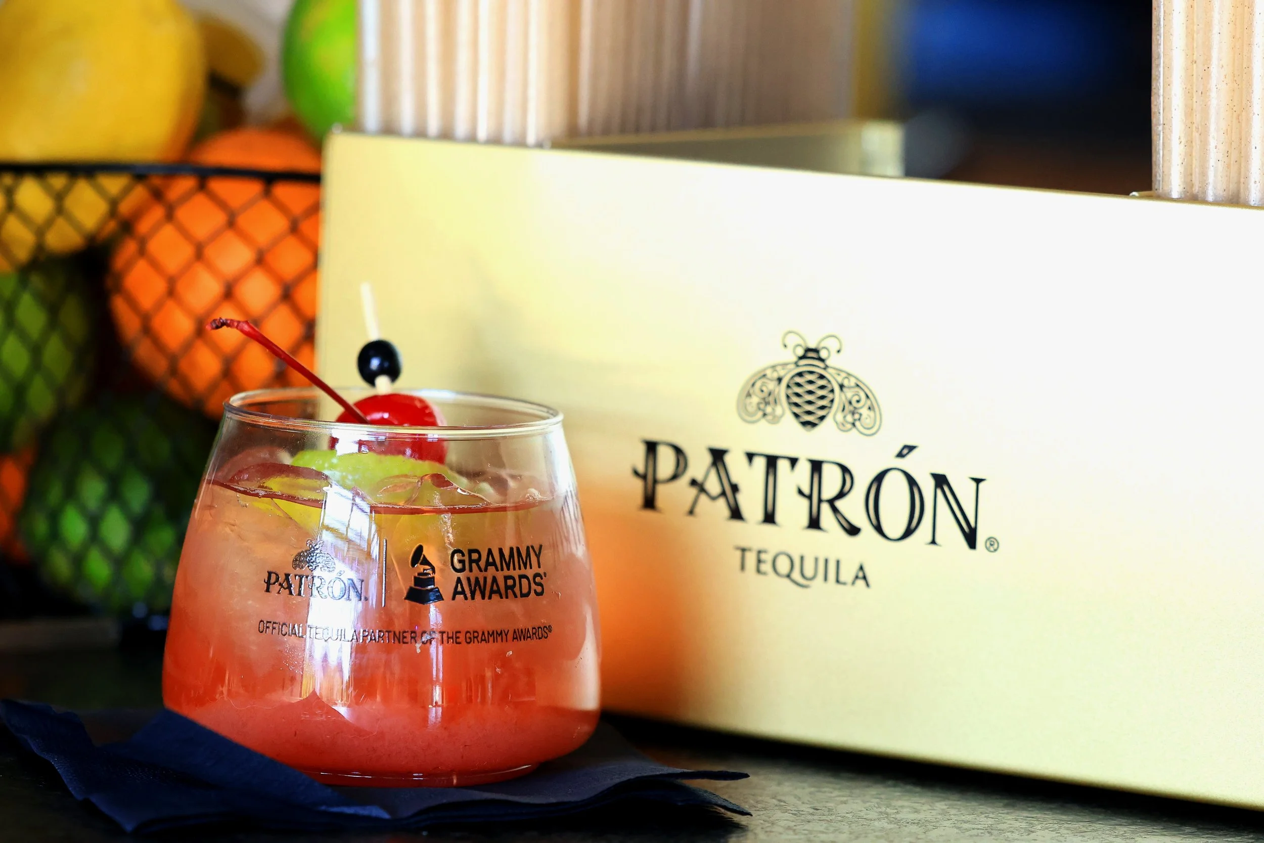 A tequila cocktail with ice, cherry, and lime slices in a glass on a black cloth, with a Patrón tequila box in the background.
