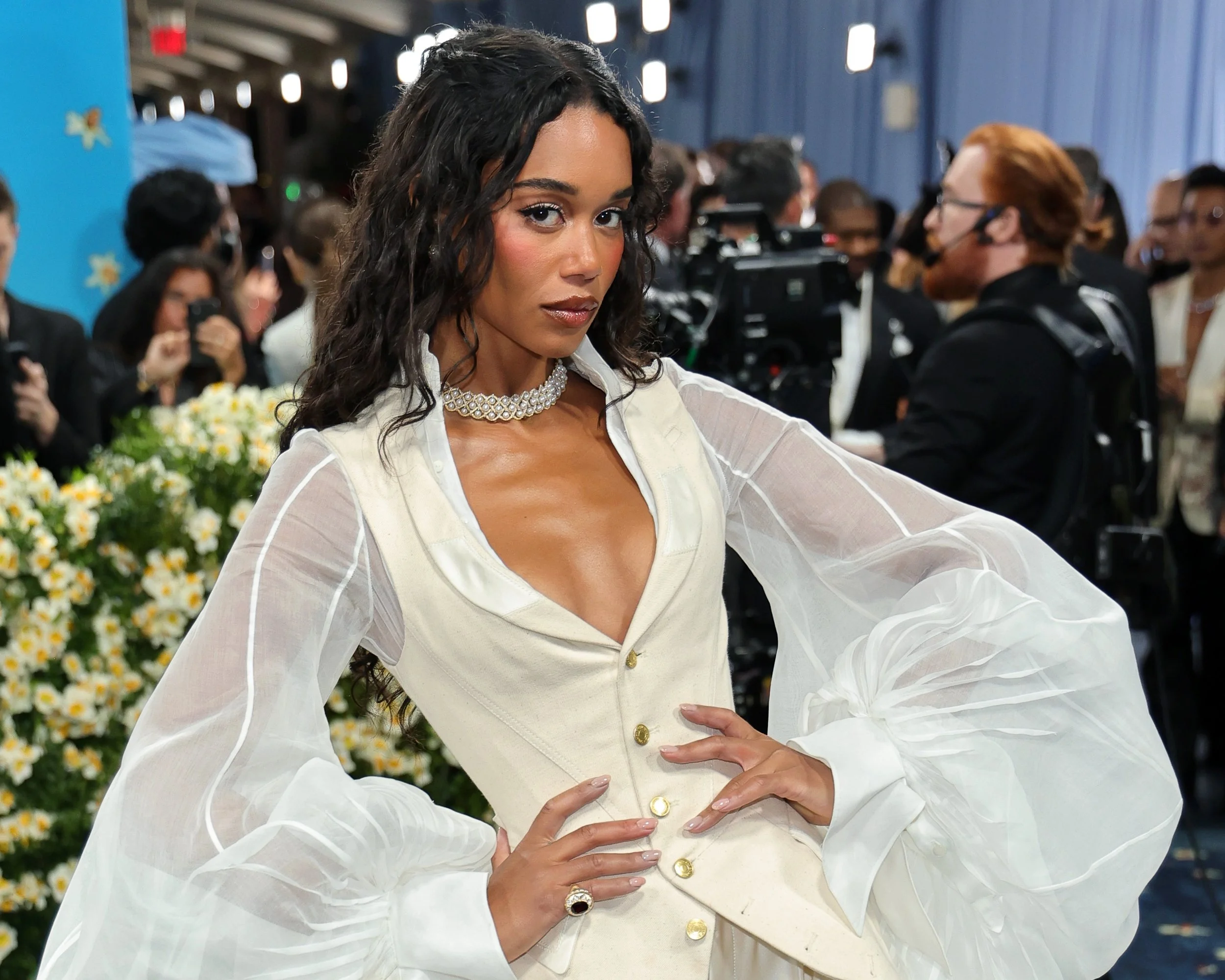 A woman with long dark curly hair wearing a white vintage-style dress with sheer puffed sleeves and gold buttons, standing in front of a crowd and photographers at an event.