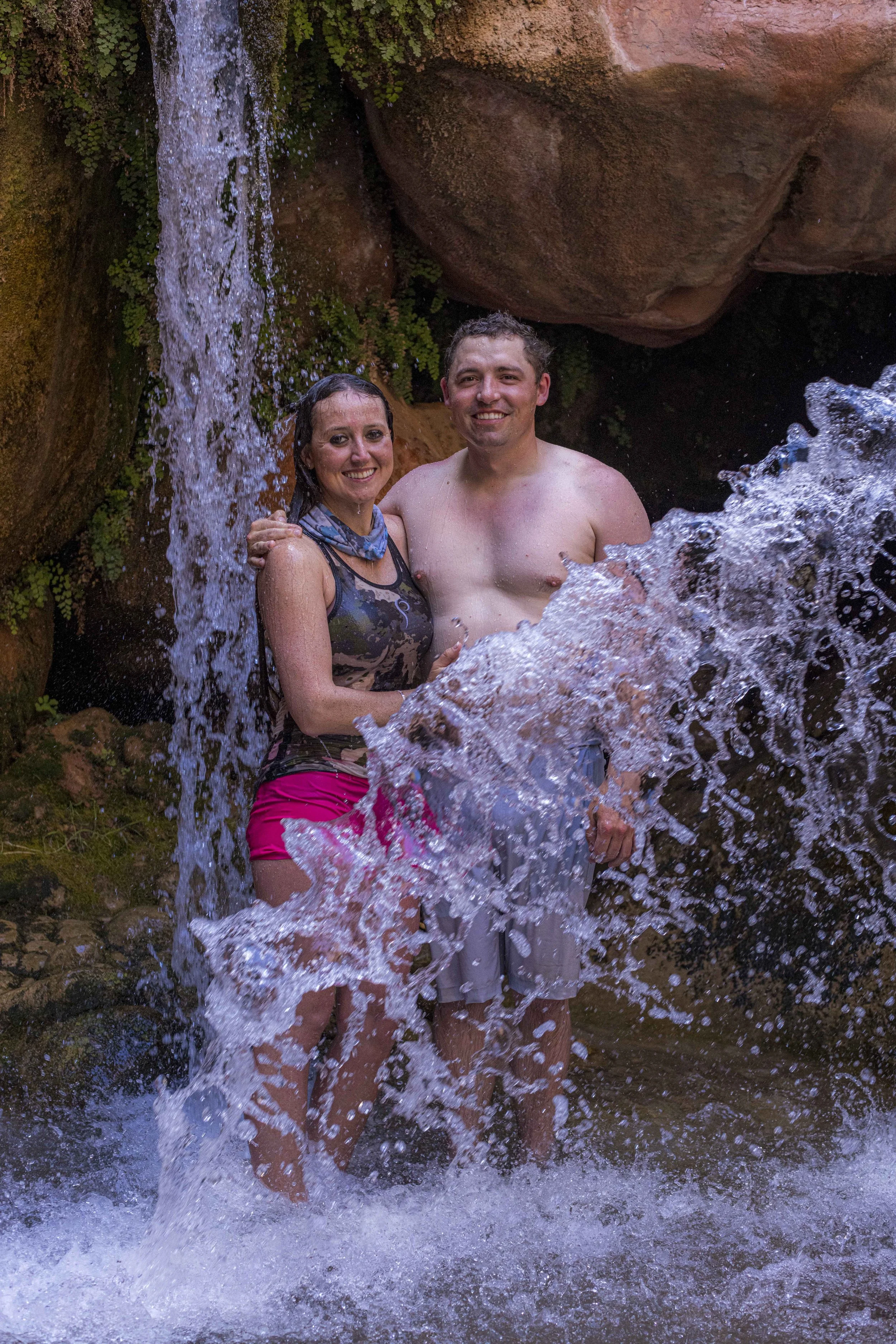 A smiling woman and man are standing under a small waterfall in a rocky outdoor setting, with water splashing around them.