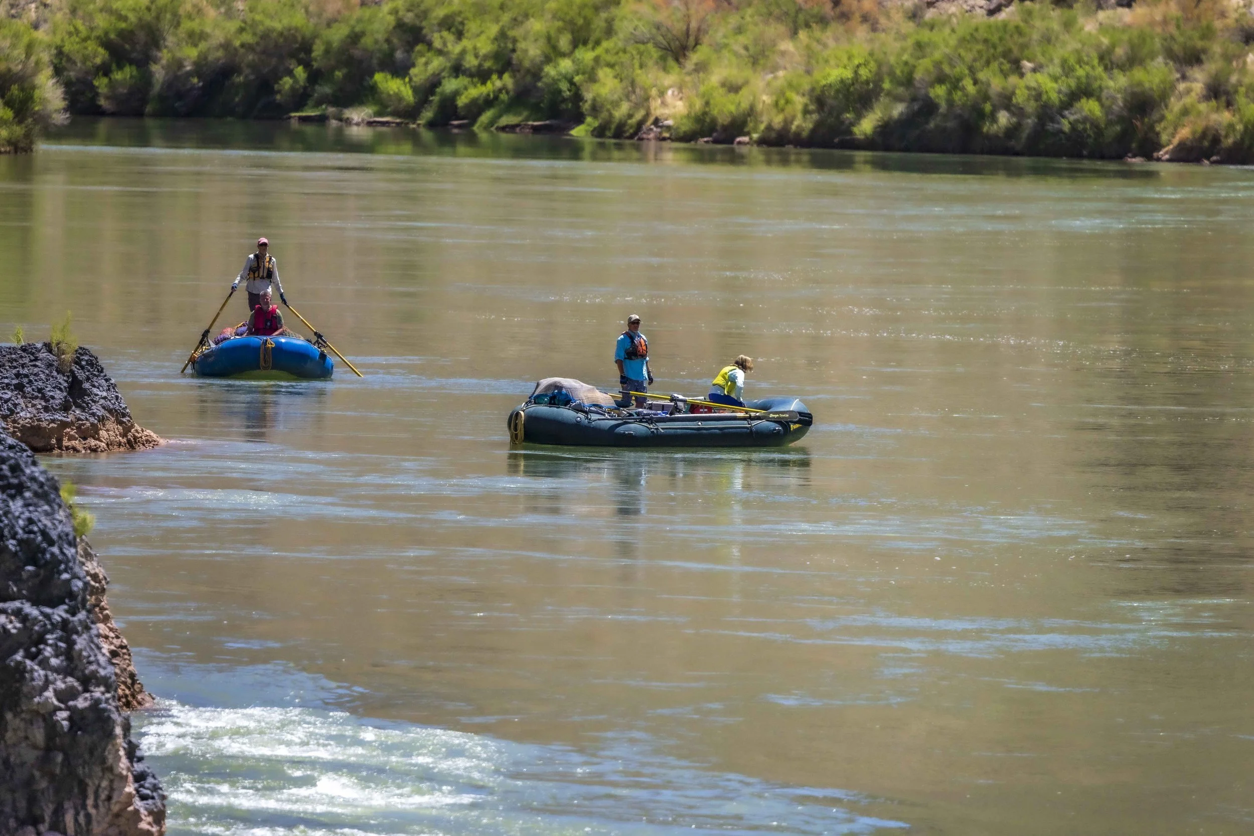 Three people in two inflatable boats on a river surrounded by green foliage.
