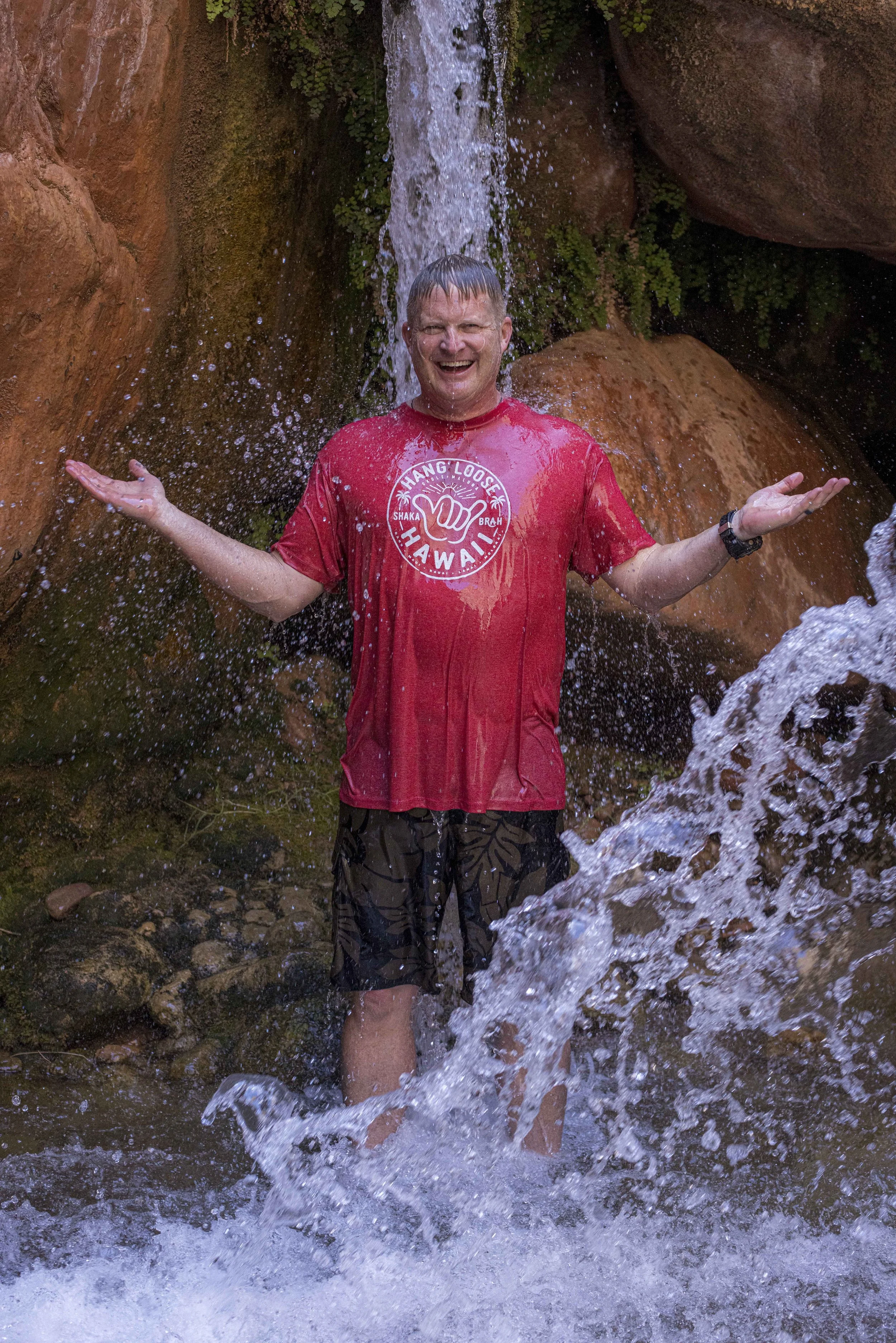 A smiling man standing under a small waterfall in a rocky, outdoor setting, with water splashing around him and his arms outstretched.