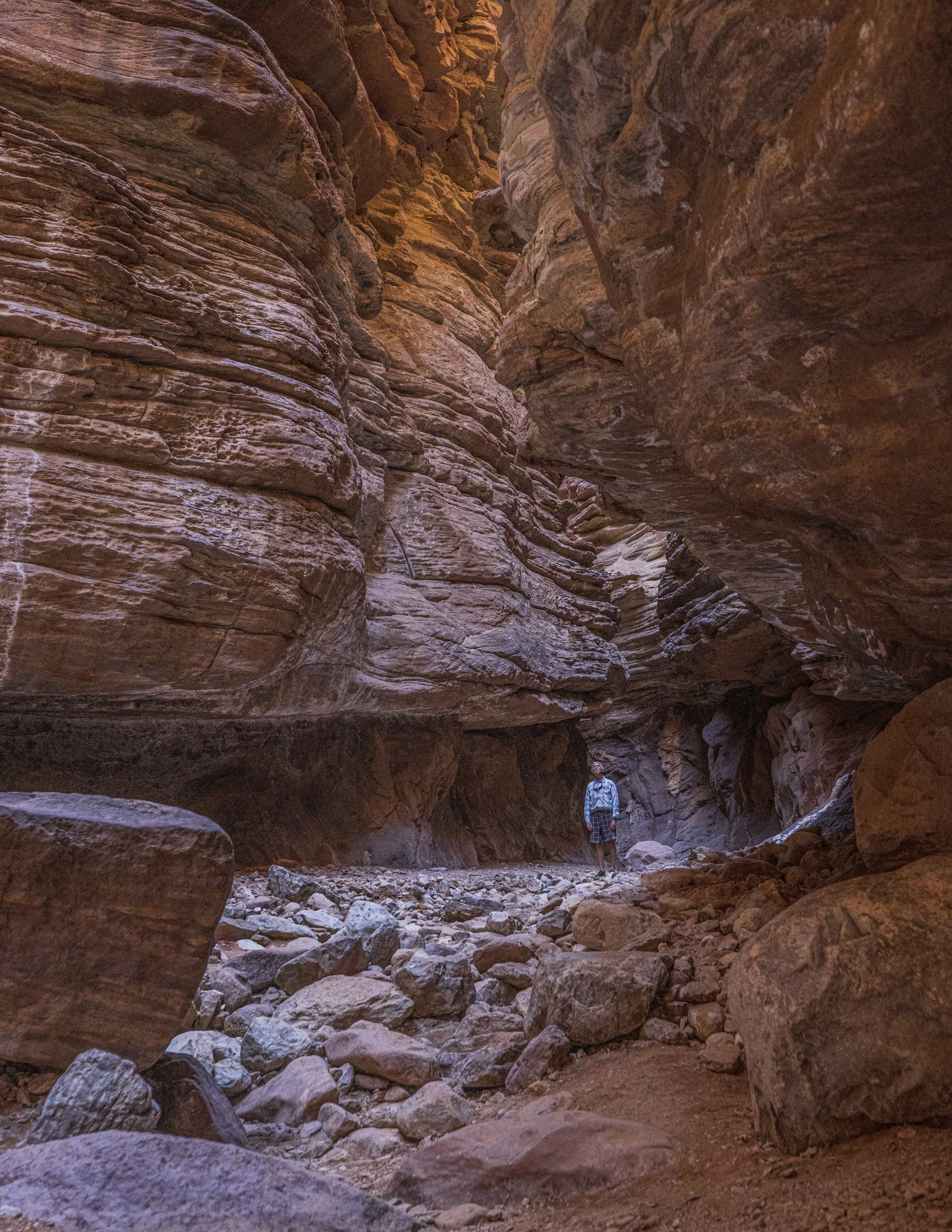A person standing inside a narrow canyon with towering red rock walls and scattered boulders on the ground.