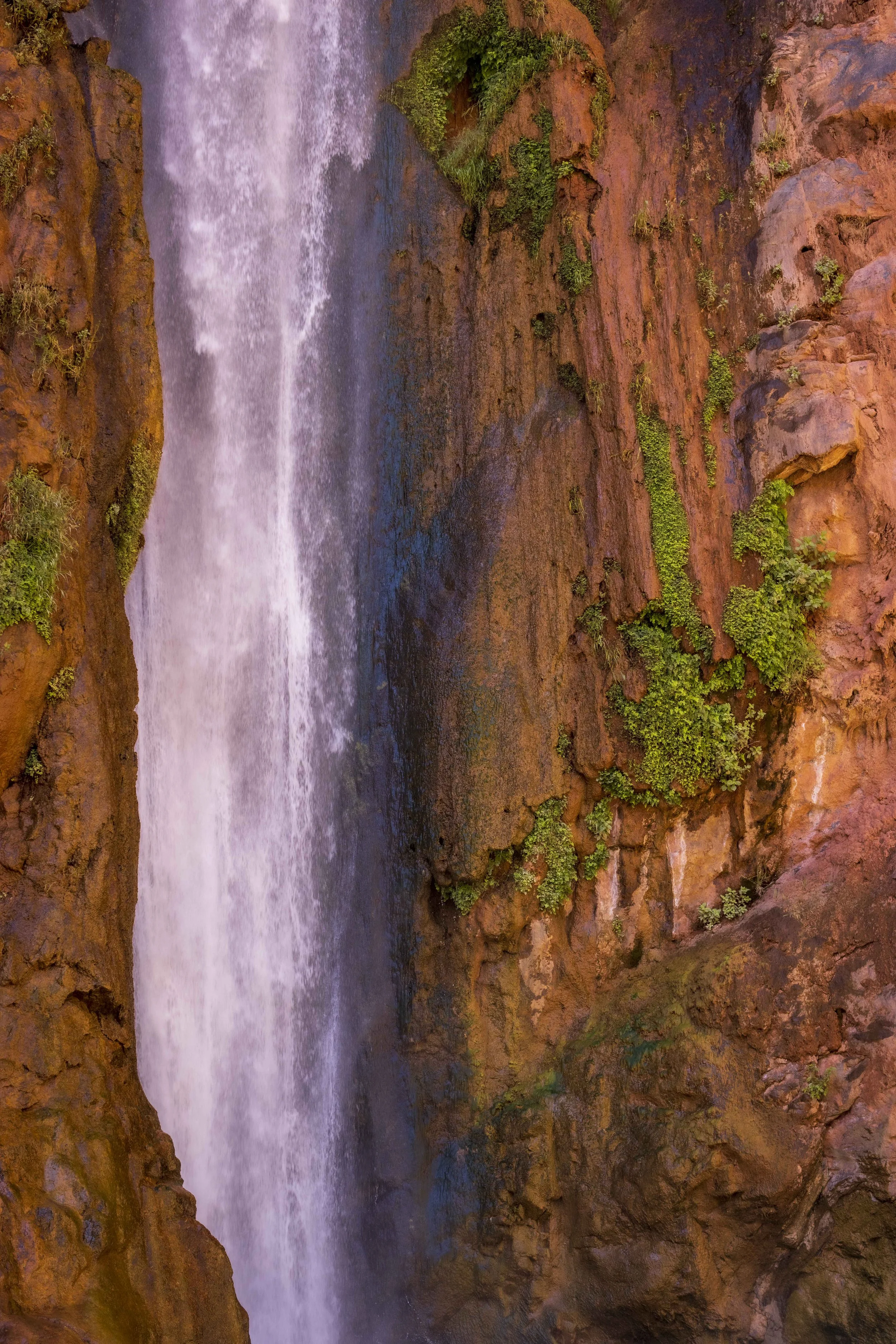 A waterfall cascading down a reddish-brown rocky cliff with green vegetation growing on the rocks.