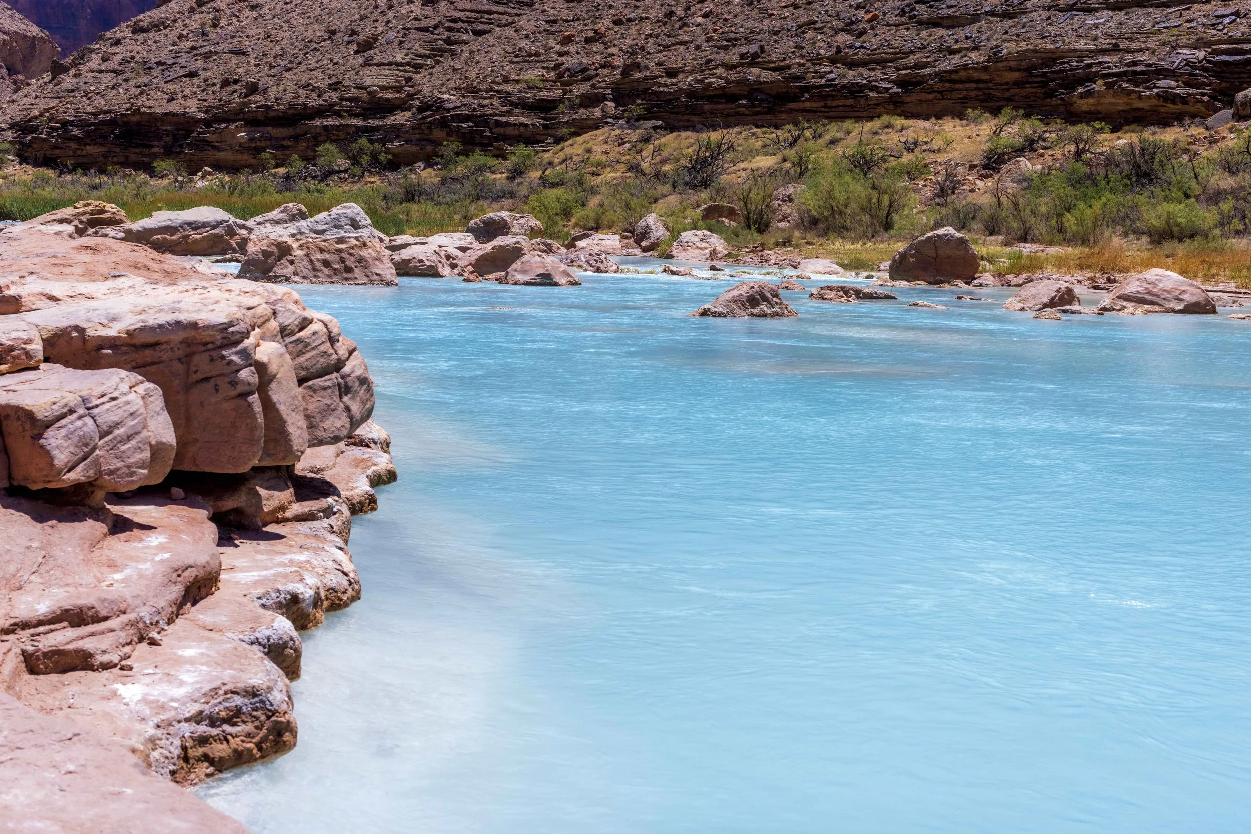 Clear blue river flowing through a desert landscape with large rocks and sparse green vegetation, surrounded by brown rocky hills.