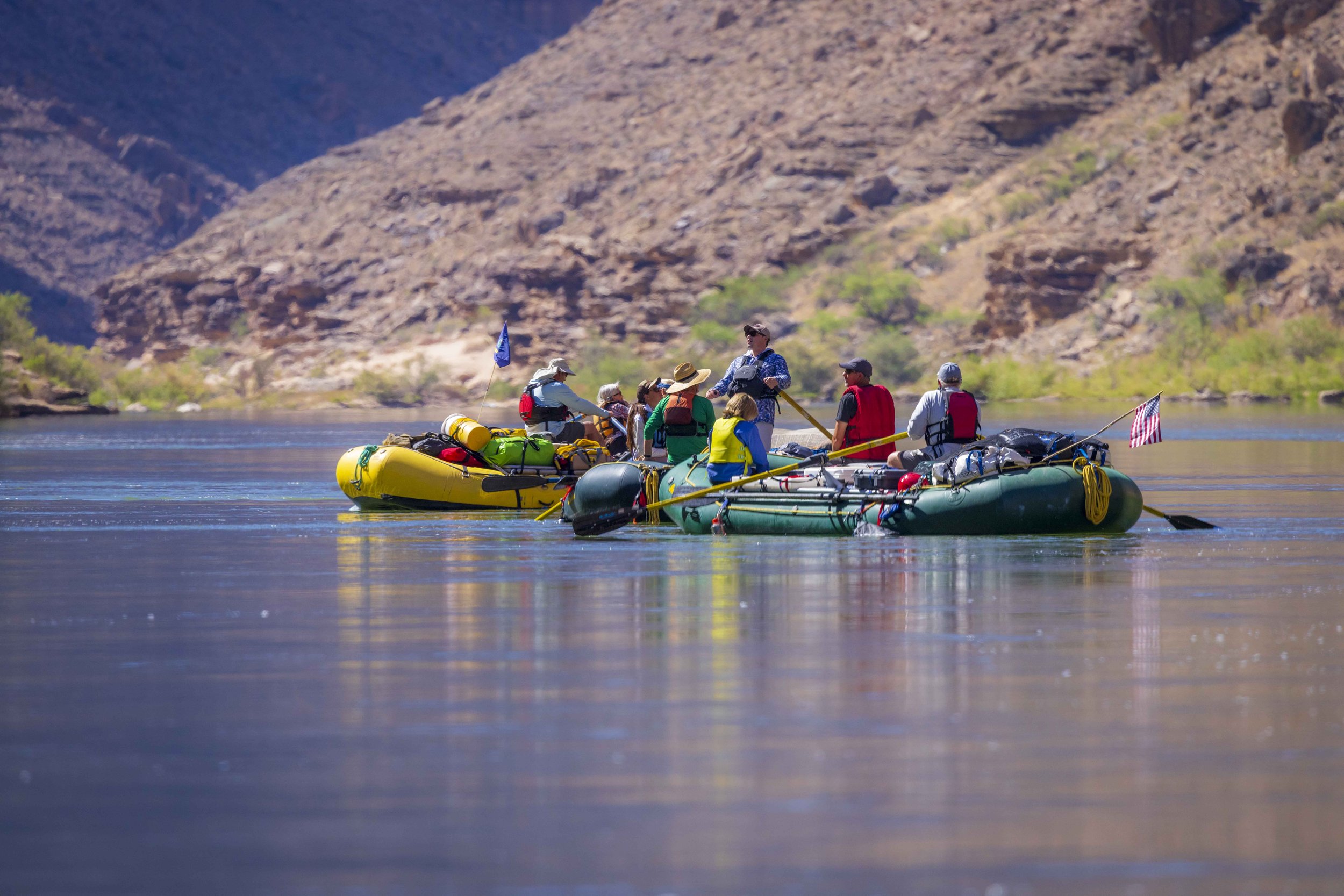 A group of people on two inflatable rafts with gear, floating on a calm river surrounded by desert mountains, during daytime.