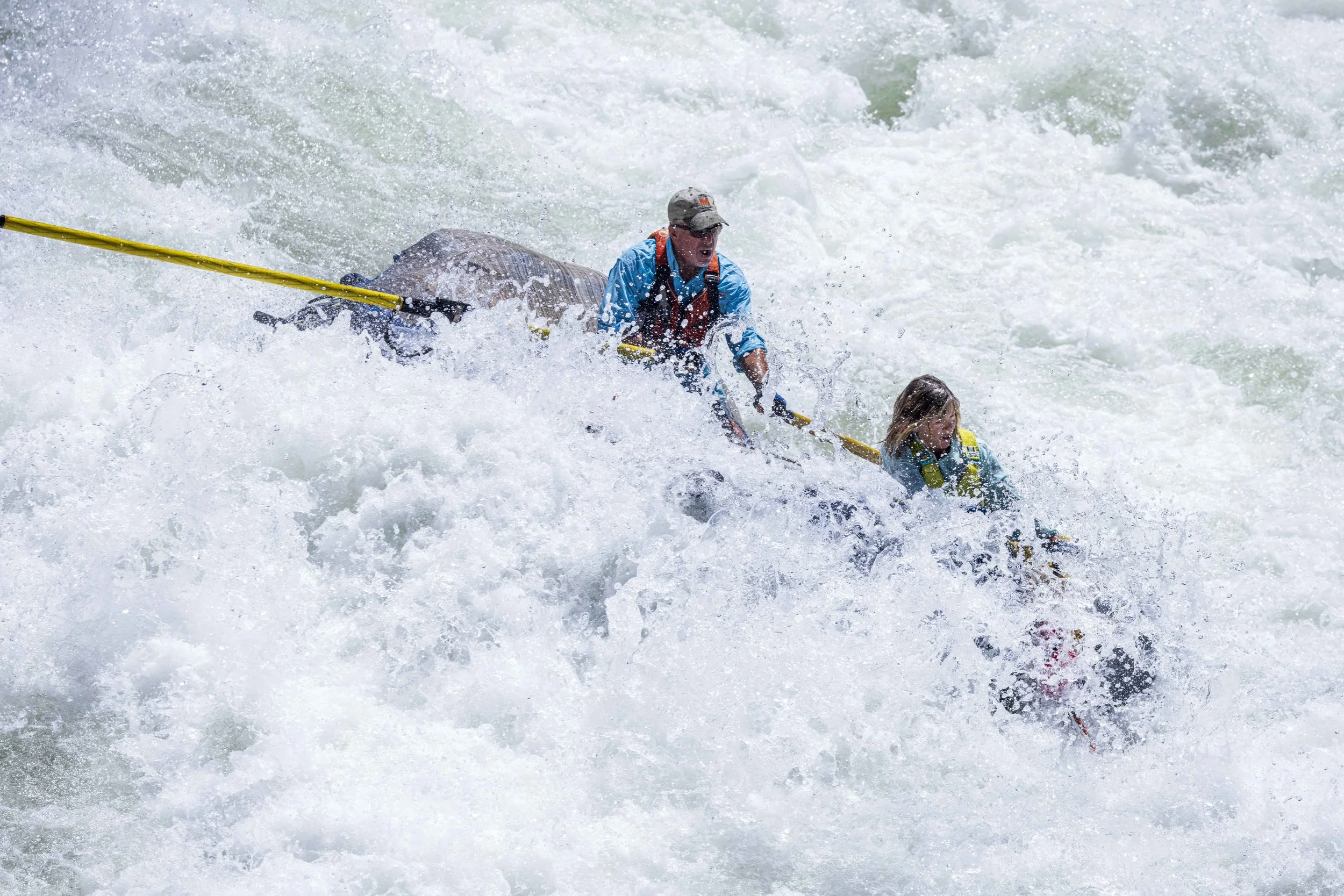 Two people white water rafting through rapids, one adult male and one young girl, both wearing life jackets and holding paddles.