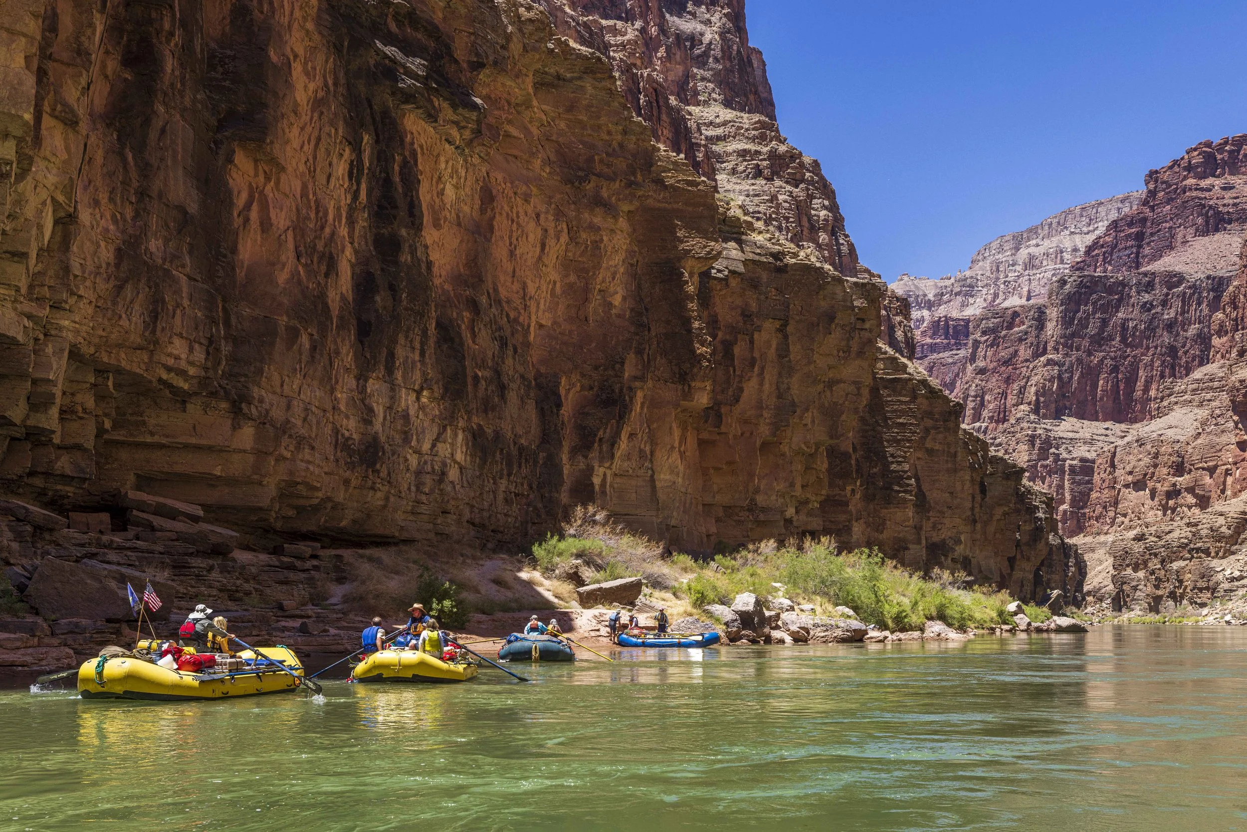 People in yellow inflatable rafts paddling on a river through a deep canyon with reddish-brown rock walls and a clear blue sky.