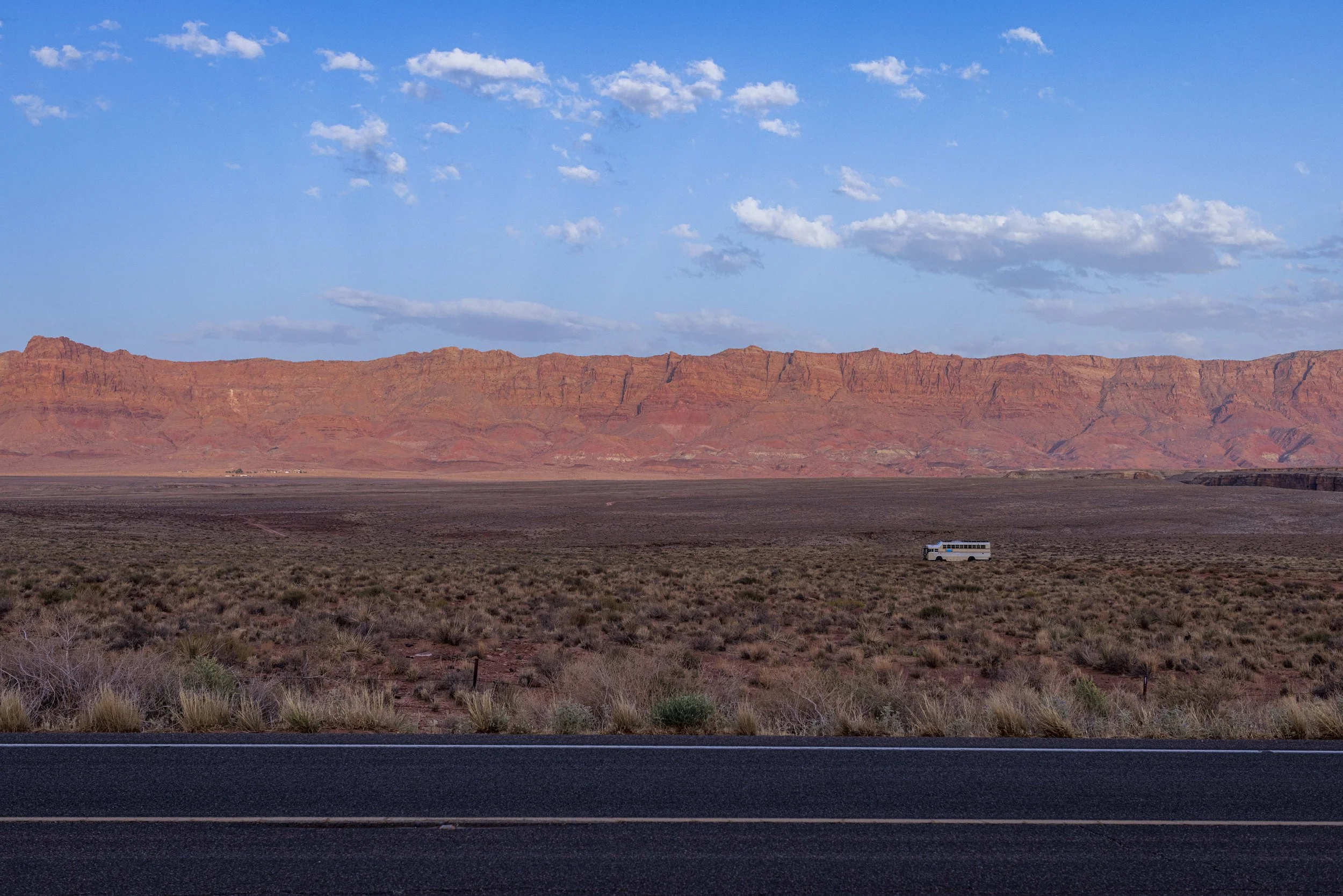 A vast desert landscape with a red mountain range in the distance, a clear blue sky with a few clouds, and a white vehicle traveling on a two-lane road in the foreground.