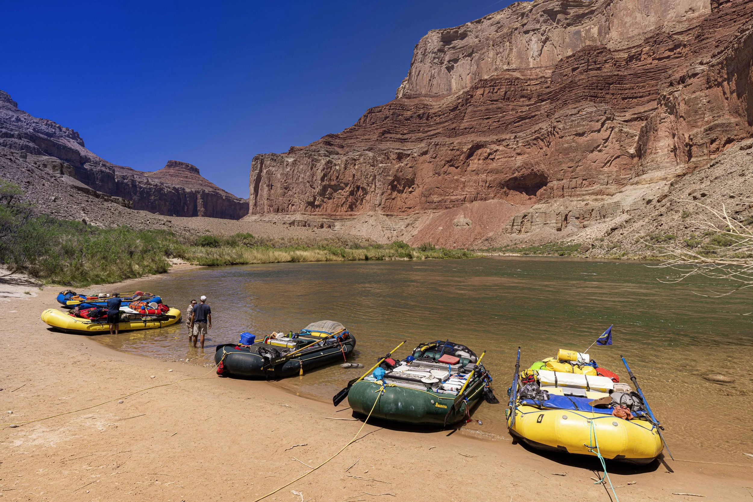 Four inflatable rafts with gear on the sandy shore of a river surrounded by tall red canyon walls and desert vegetation under a clear blue sky.