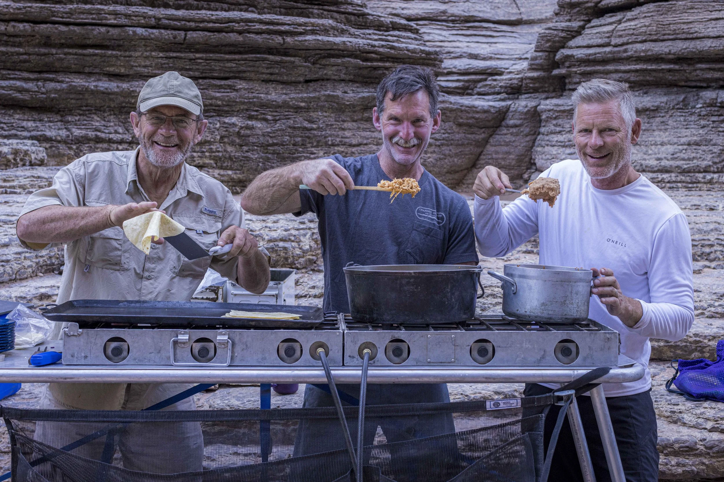 Three men smiling and cooking outdoors in front of a rocky cliff, with one preparing tortilla, another holding a spoon of food, and the third holding a spoon of fried chicken.