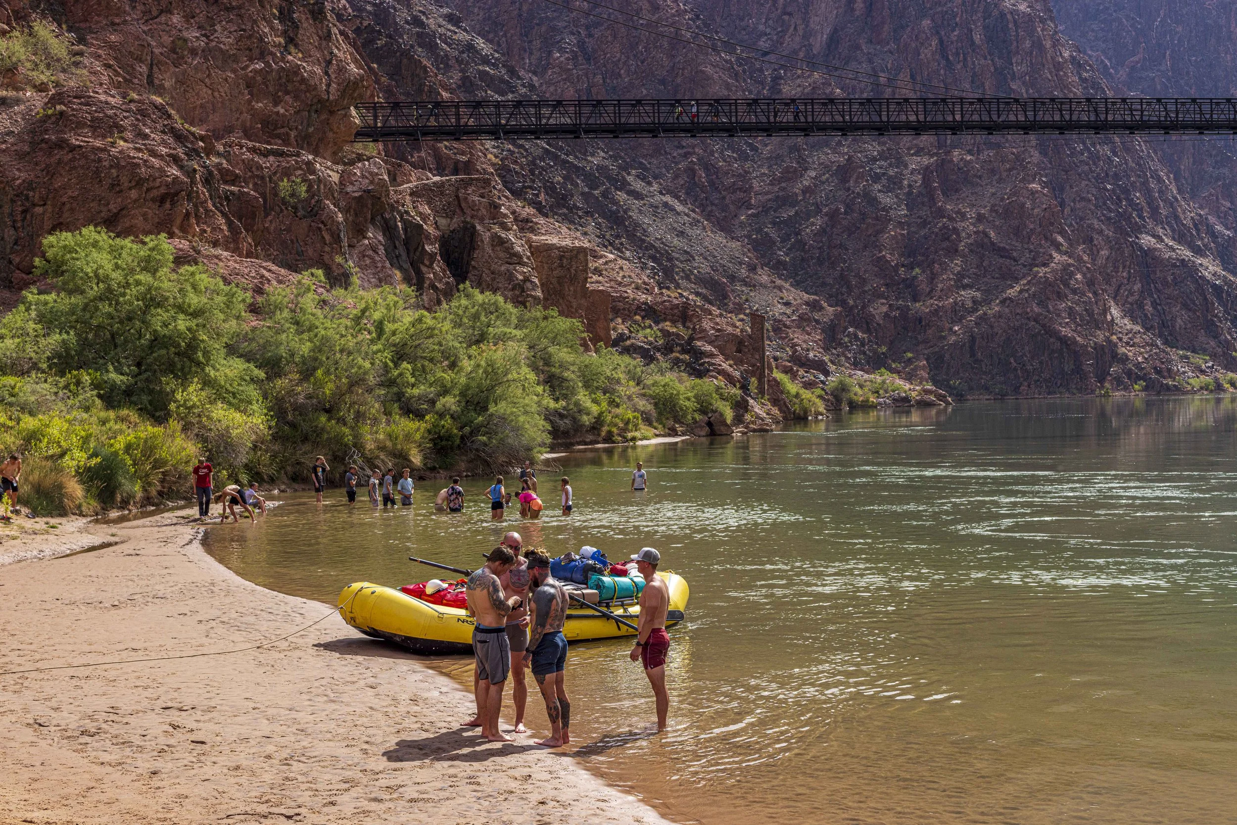 People standing on a beach near a yellow inflatable boat, with some in the water and others on the shore, surrounded by green trees and rocky cliffs, and a bridge overhead.