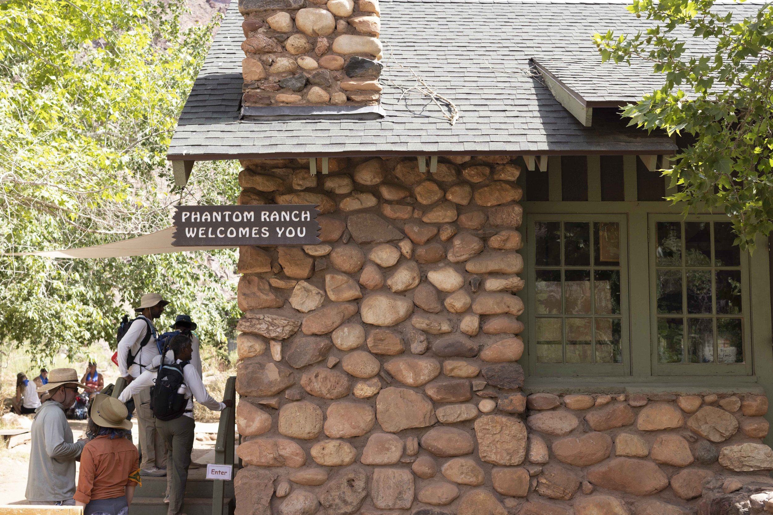 People standing outside a stone building with a sign that reads "Phantom Ranch Welcomes You". There are trees around and a small group of individuals, some with backpacks and hats, are entering or waiting to enter the building.