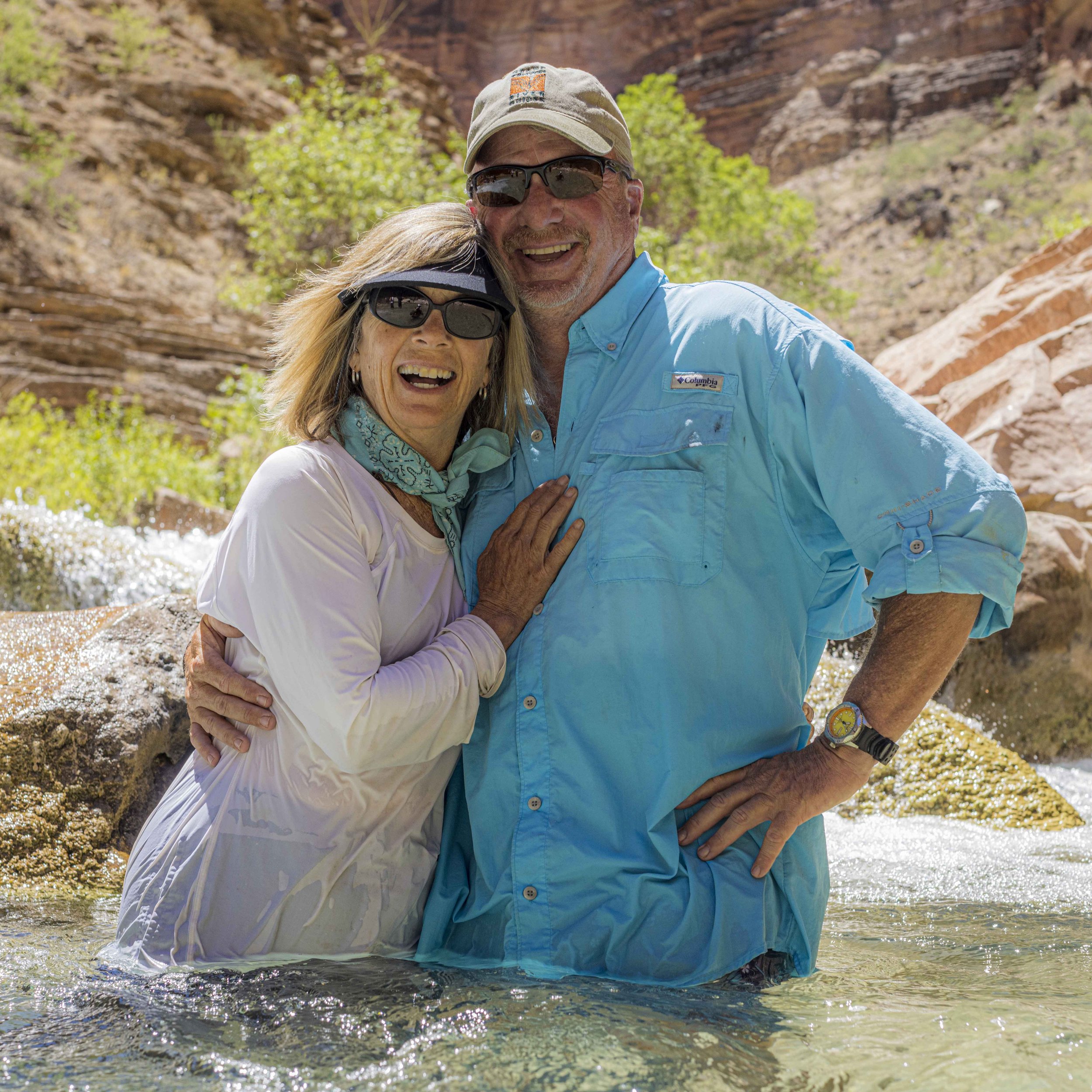 A happy couple stands in a river with rocky and green landscape in the background, both wearing outdoor clothing and sunglasses, smiling at the camera.