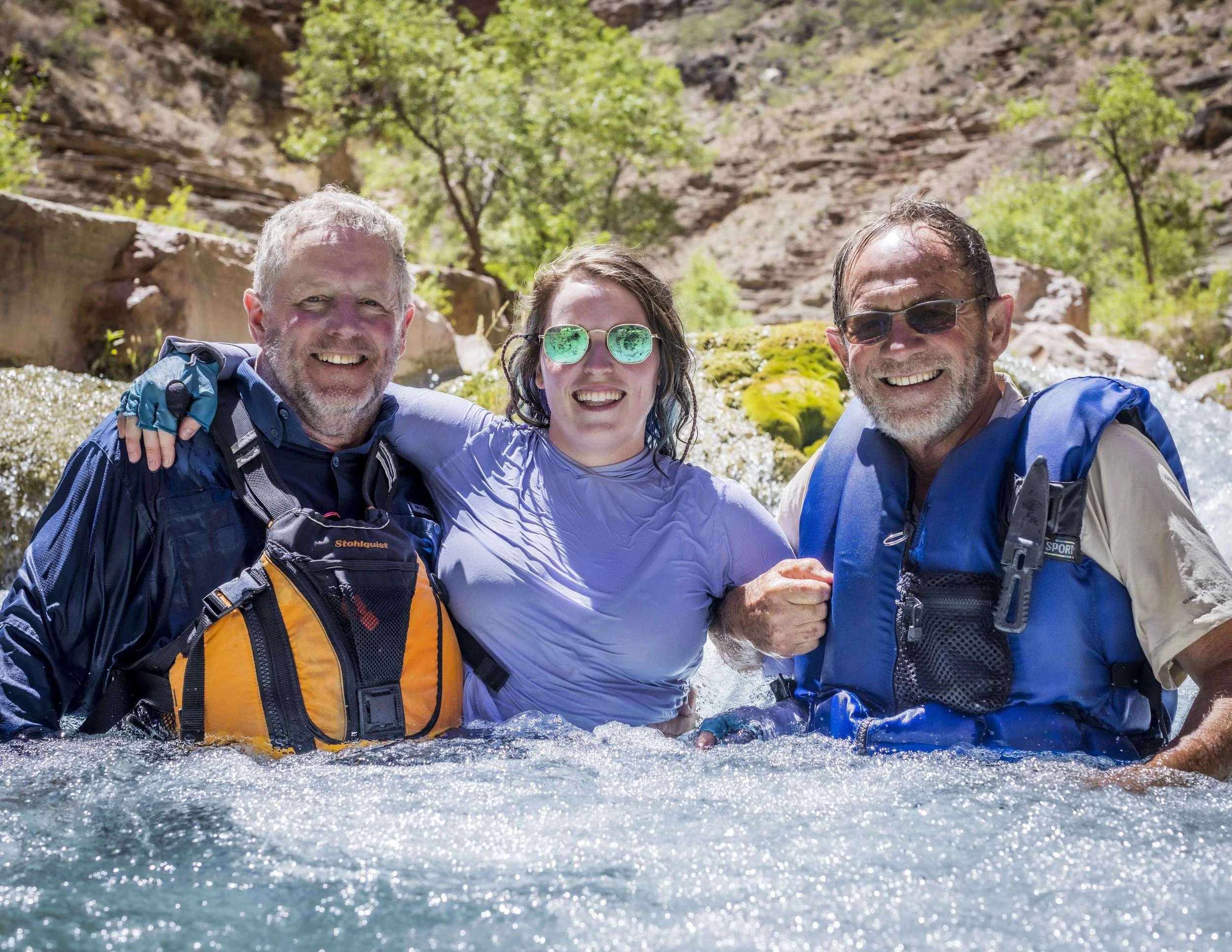 Three people smiling in a river, wearing outdoor gear and sunglasses, with rocky cliffs and greenery in the background.