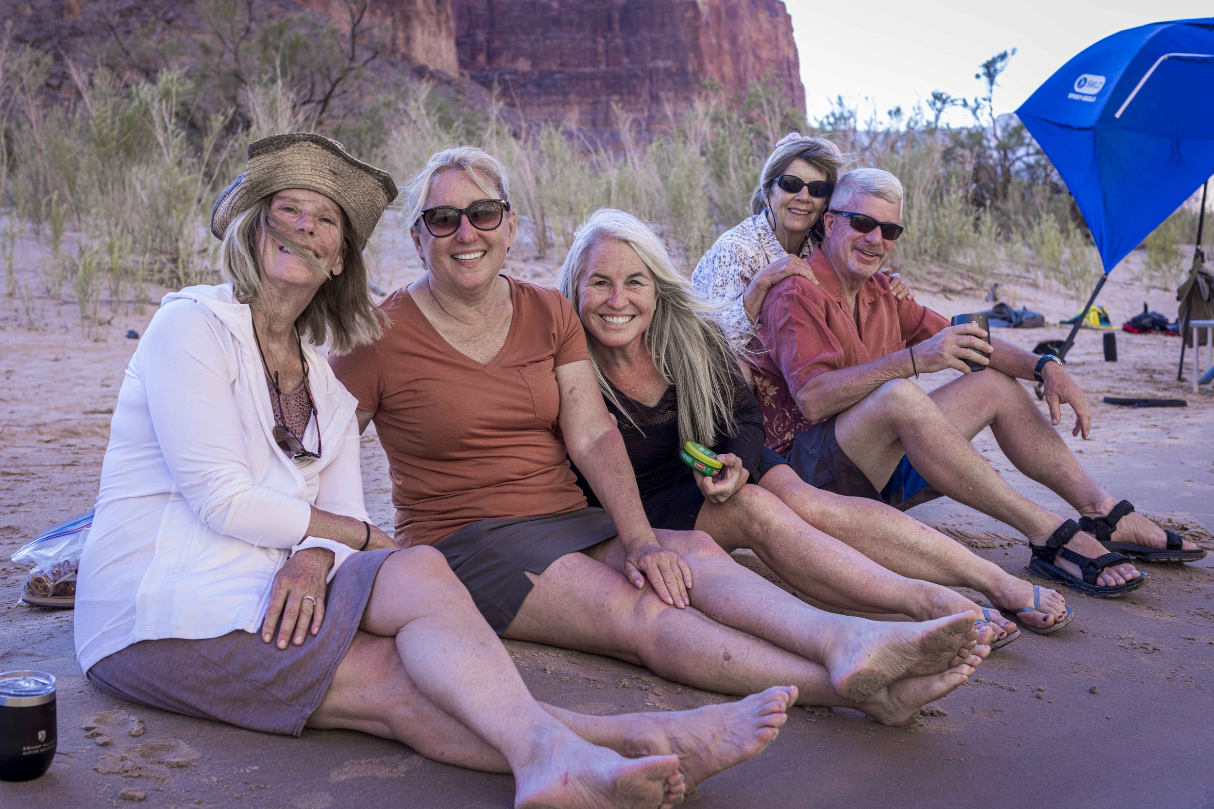 Five smiling older adults sitting on the sand at a beach, enjoying a casual outdoor gathering, with a large blue beach umbrella and rocky cliffs in the background.