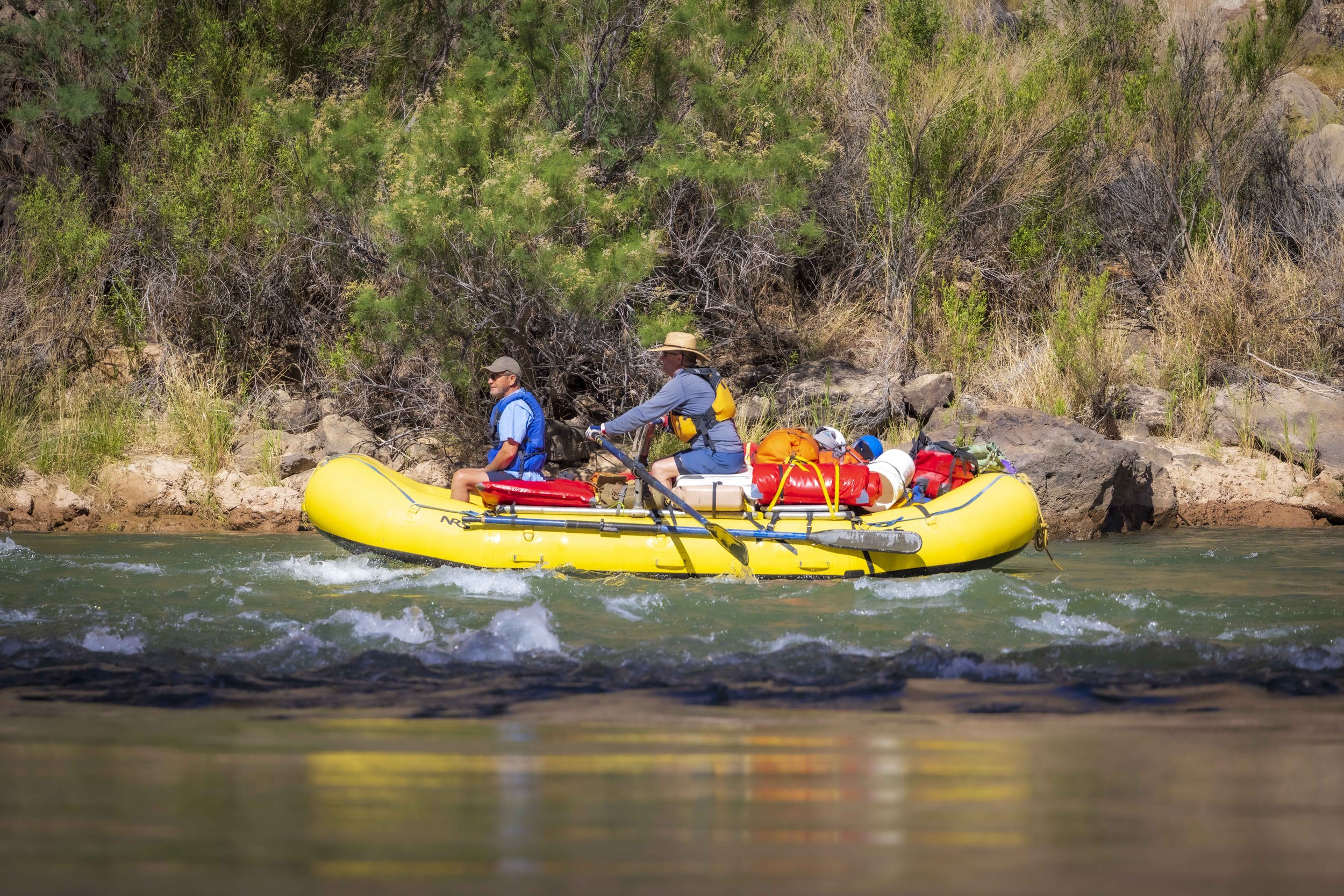 Two people in yellow inflatable raft on a river, surrounded by green trees and rocks.