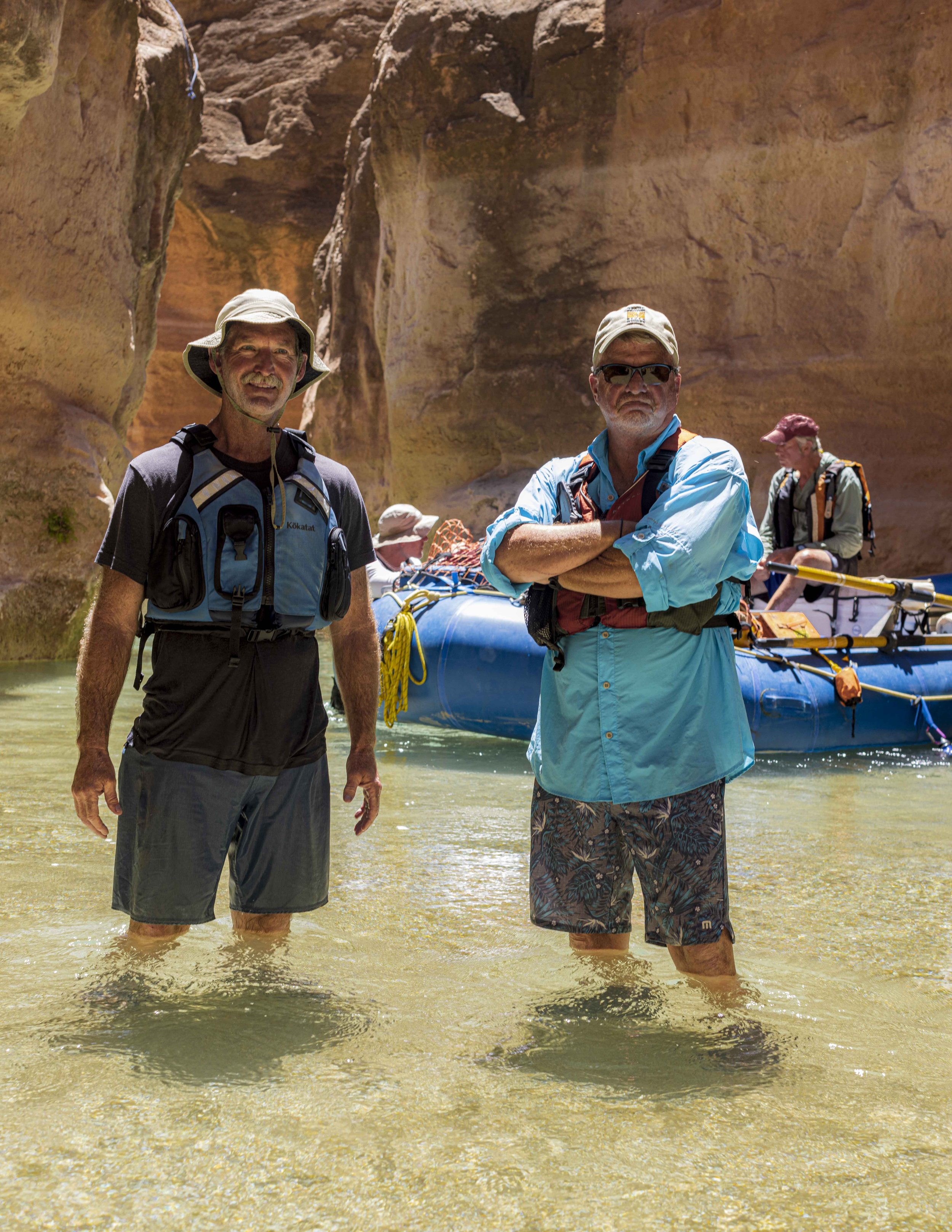 Two men standing in shallow water with a canyon and a raft in the background.
