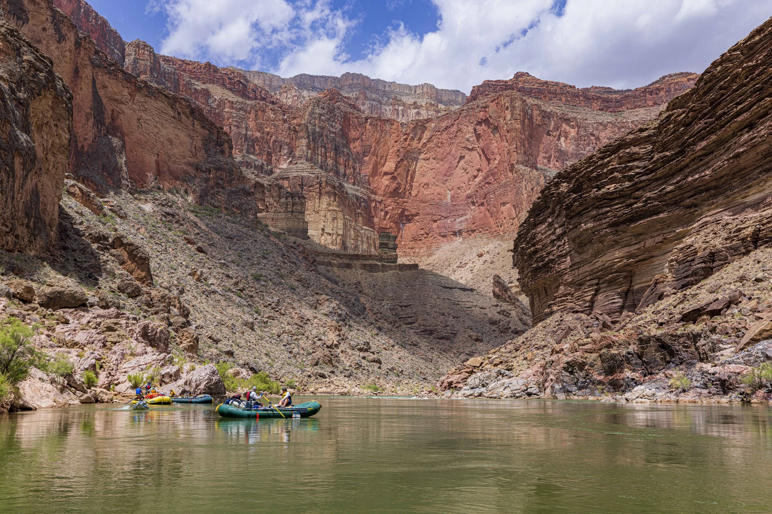 Rafting boats floating on the Colorado River in the Grand Canyon surrounded by towering red and brown rock formations and a partly cloudy sky.