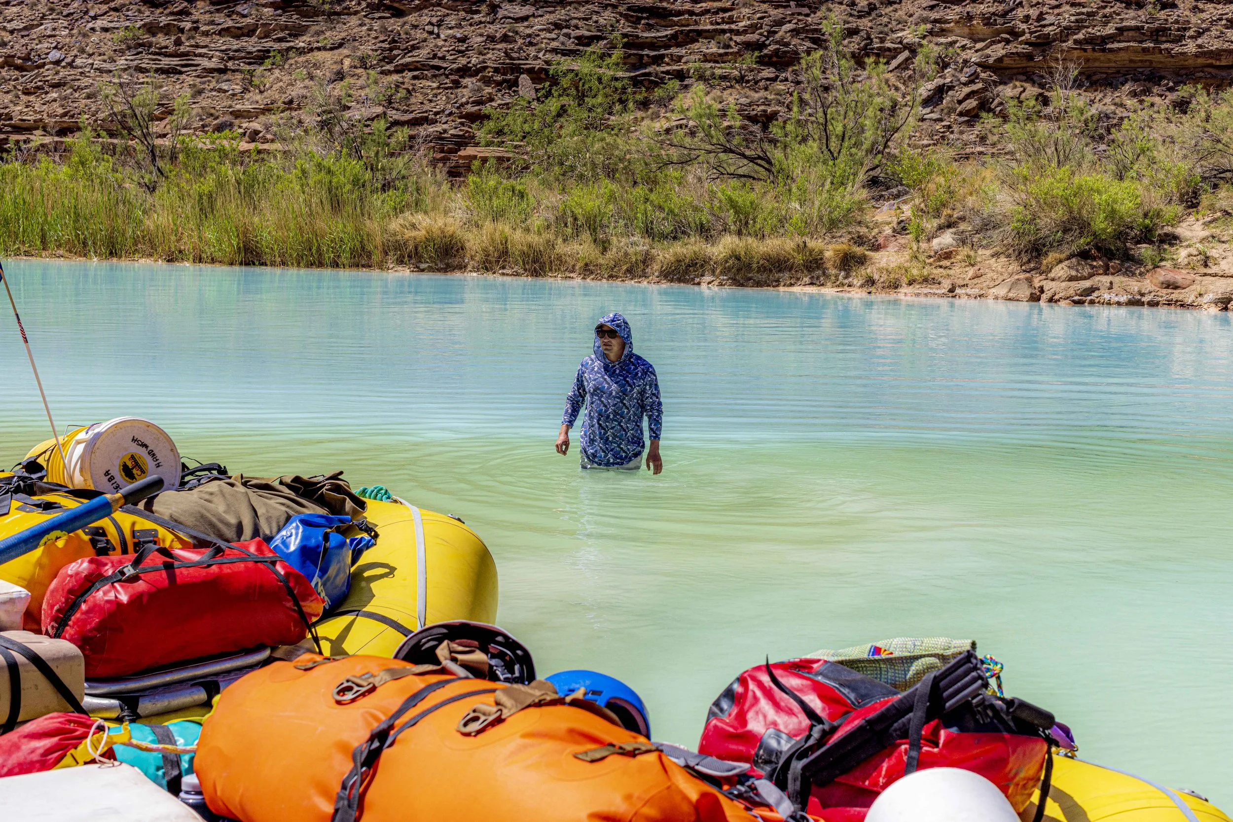 A person wearing a blue patterned rain jacket and sunglasses wading in a greenish body of water, with a yellow raft and various colorful gear and backpacks on the shore in the foreground, and rocky terrain with sparse green bushes in the background.