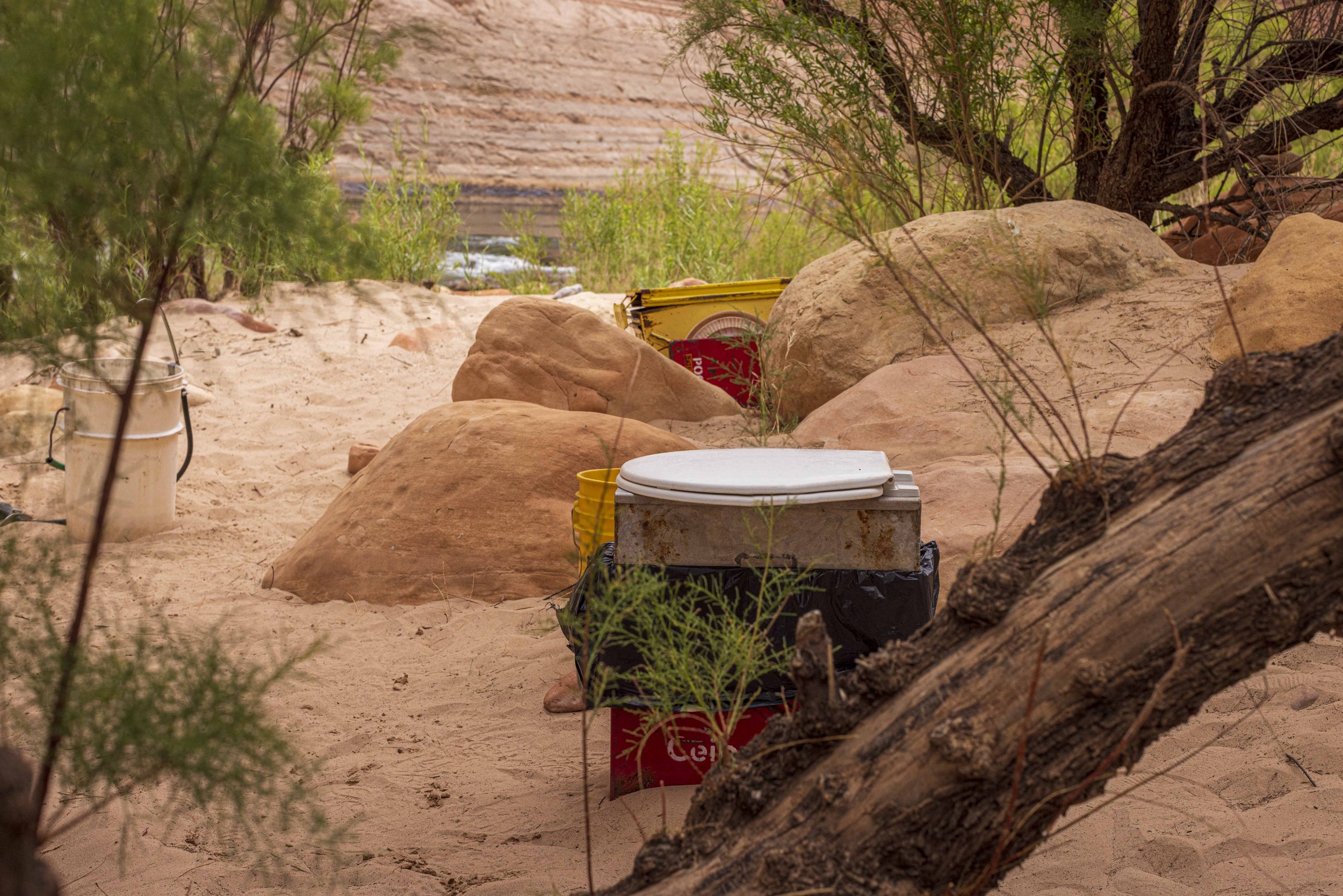 Campground with a white cooler, yellow bucket, and other trash on sandy ground surrounded by rocks and sparse green bushes.