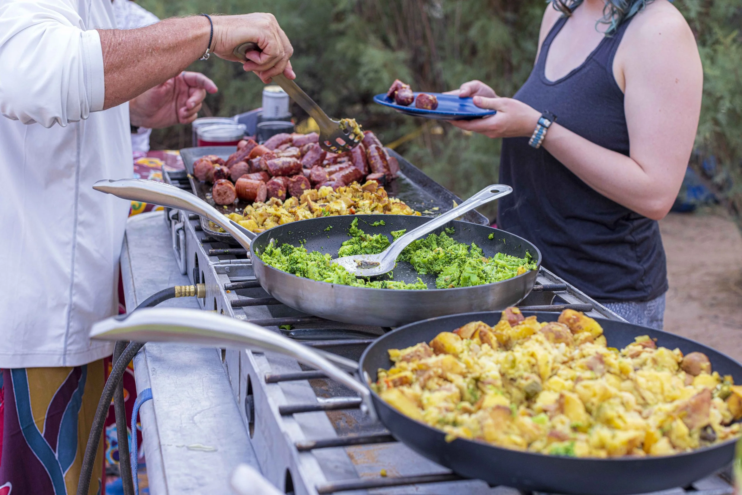 People serving food at an outdoor gathering, with various dishes including broccoli, roasted potatoes, and sausages on the table.