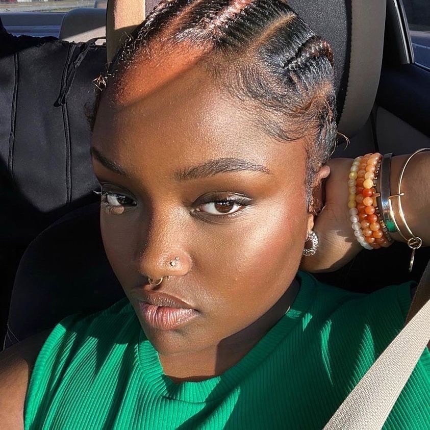 A young woman with braided hair, wearing a green top, sitting in a car, taking a selfie. She has a septum piercing, hoop earrings, and multiple bracelets on her wrist.