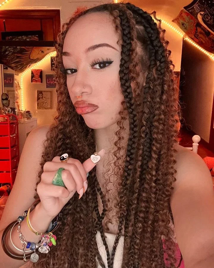 A young woman with long, curly hair styled in twists, holding her index finger to her lips. She is wearing rings, bracelets, and has a nose piercing, with makeup including eyeliner and lip gloss. The room behind her is decorated with colorful artwork