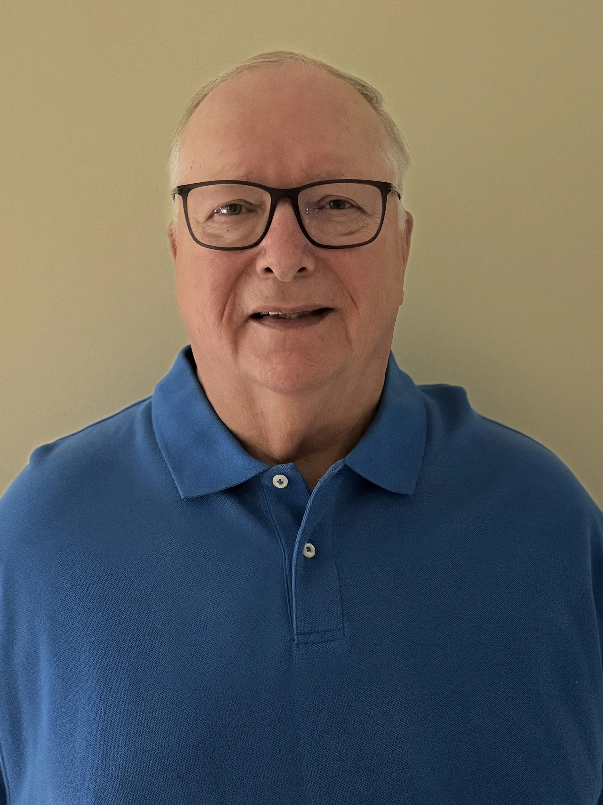 A middle-aged man with glasses and short light hair, wearing a blue polo shirt, standing against a plain yellow background.