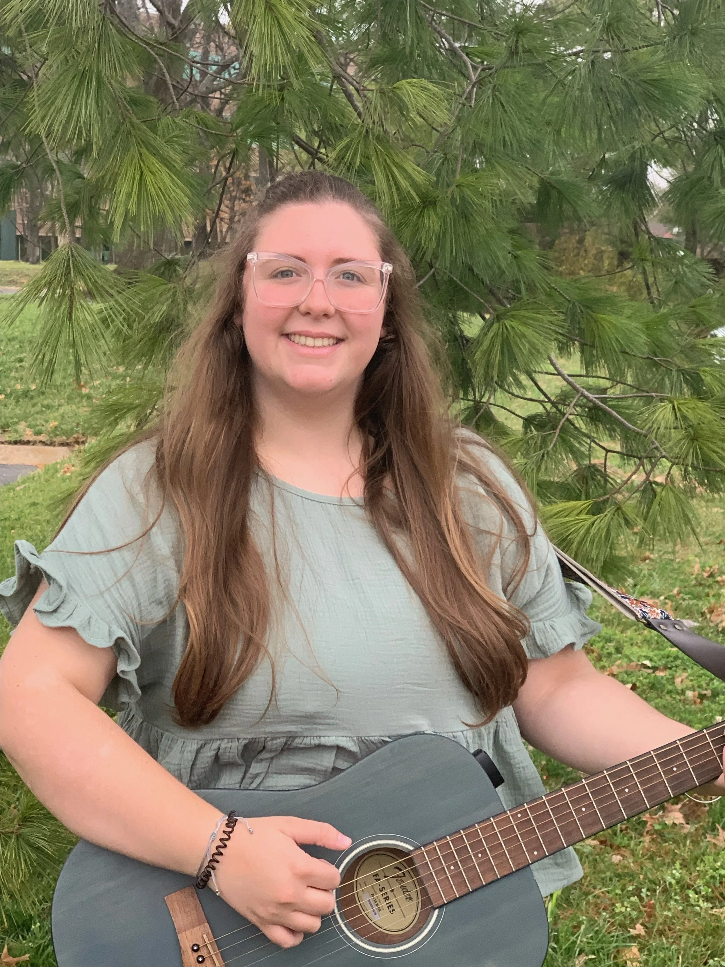 White woman with a green short sleeved shirt holding a guitar smiling in front of a tree