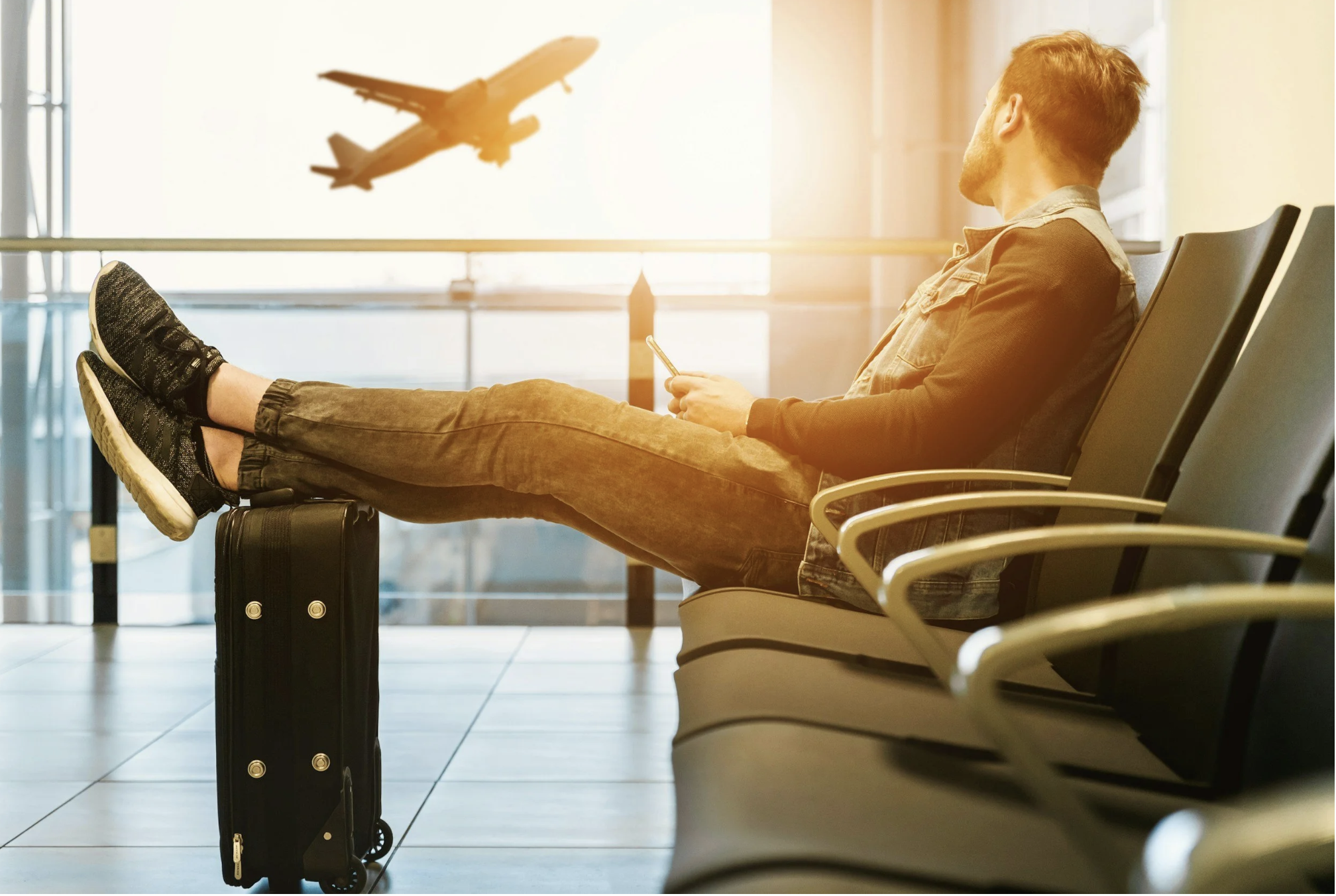 Traveler sitting in an airport terminal with luggage while watching a plane take off after preparing for an upcoming trip.
