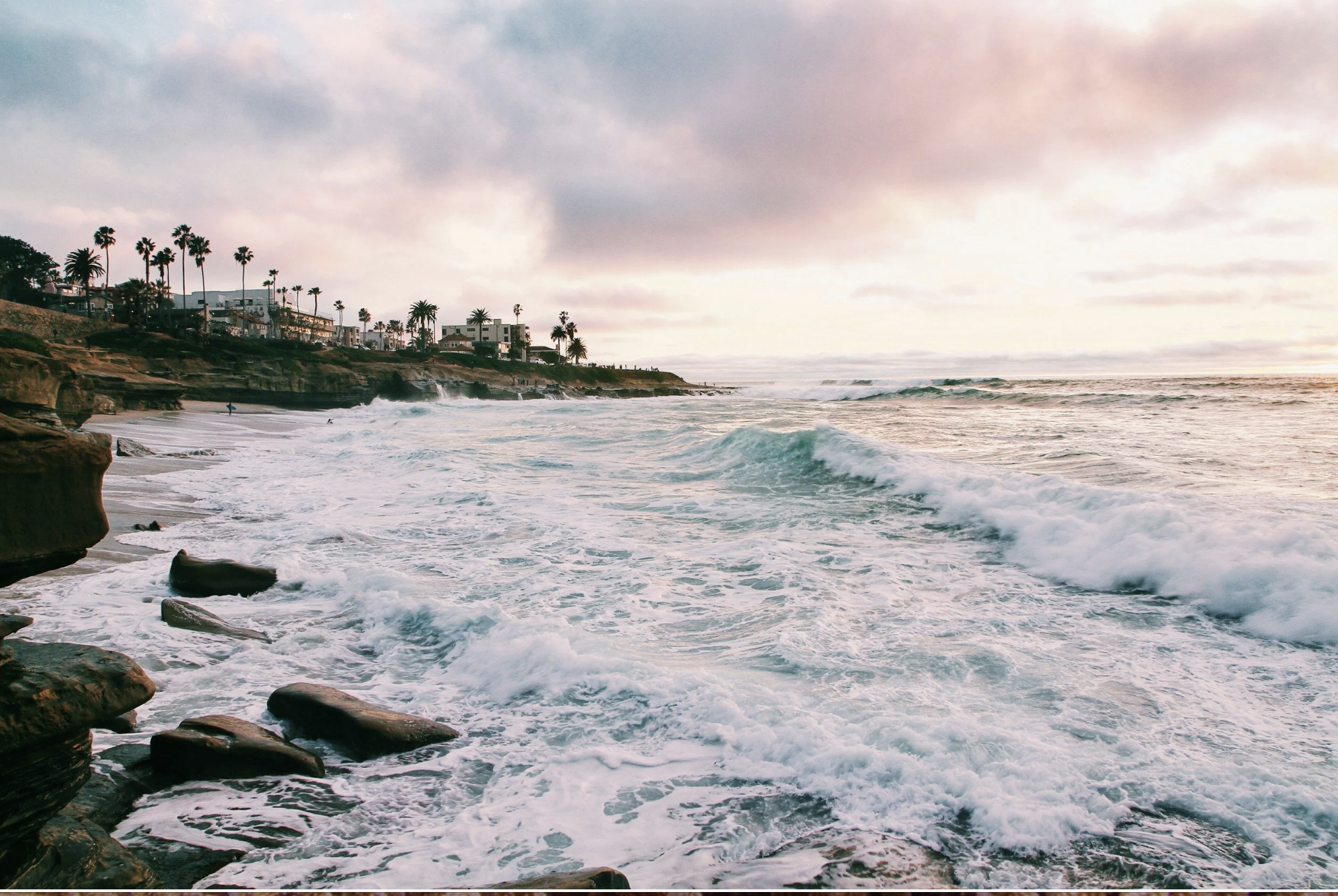 Waves crashing along a San Diego beach in California with palm trees and coastal homes at sunset.